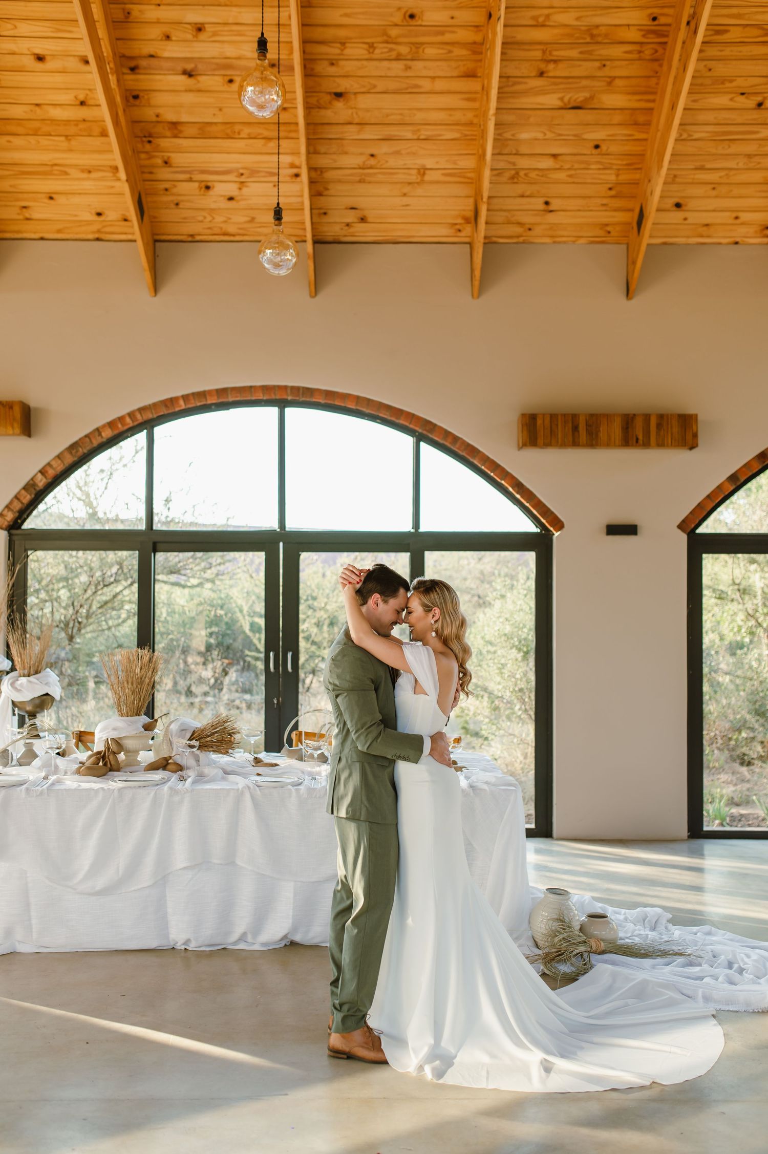 Romantic wedding couple dancing in a venue with wooden beam ceiling and arched windows.