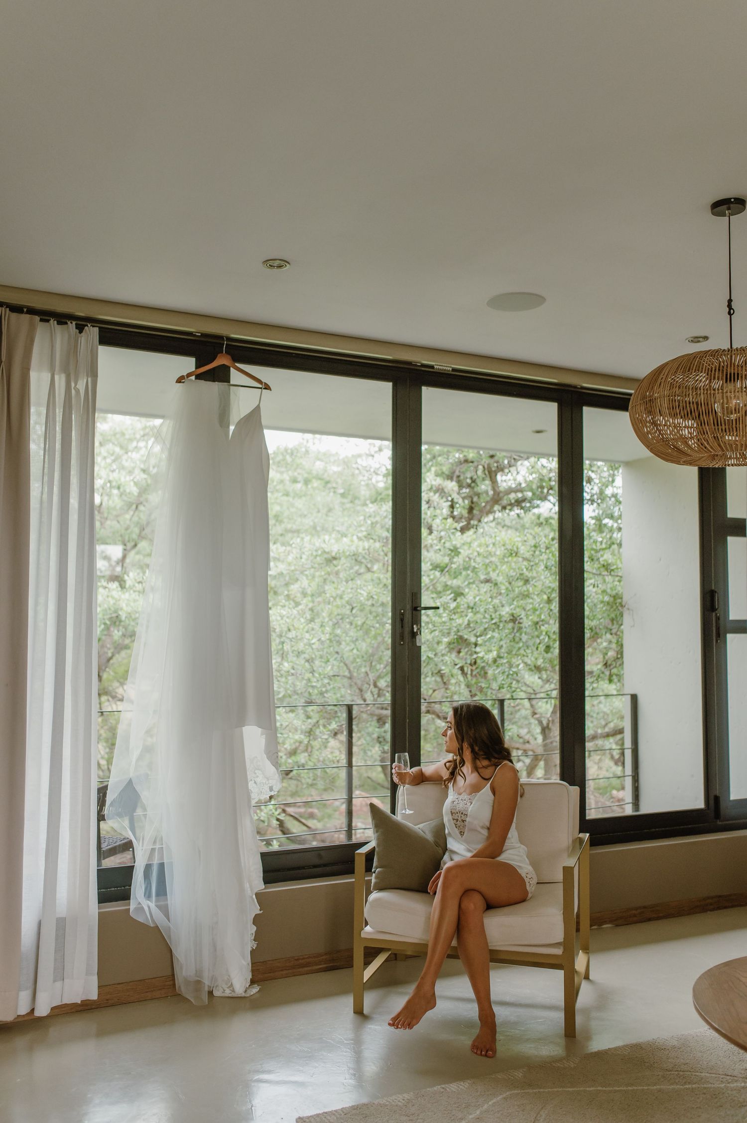 Wedding dress hangs by window as woman in white sits in modern room with natural light.