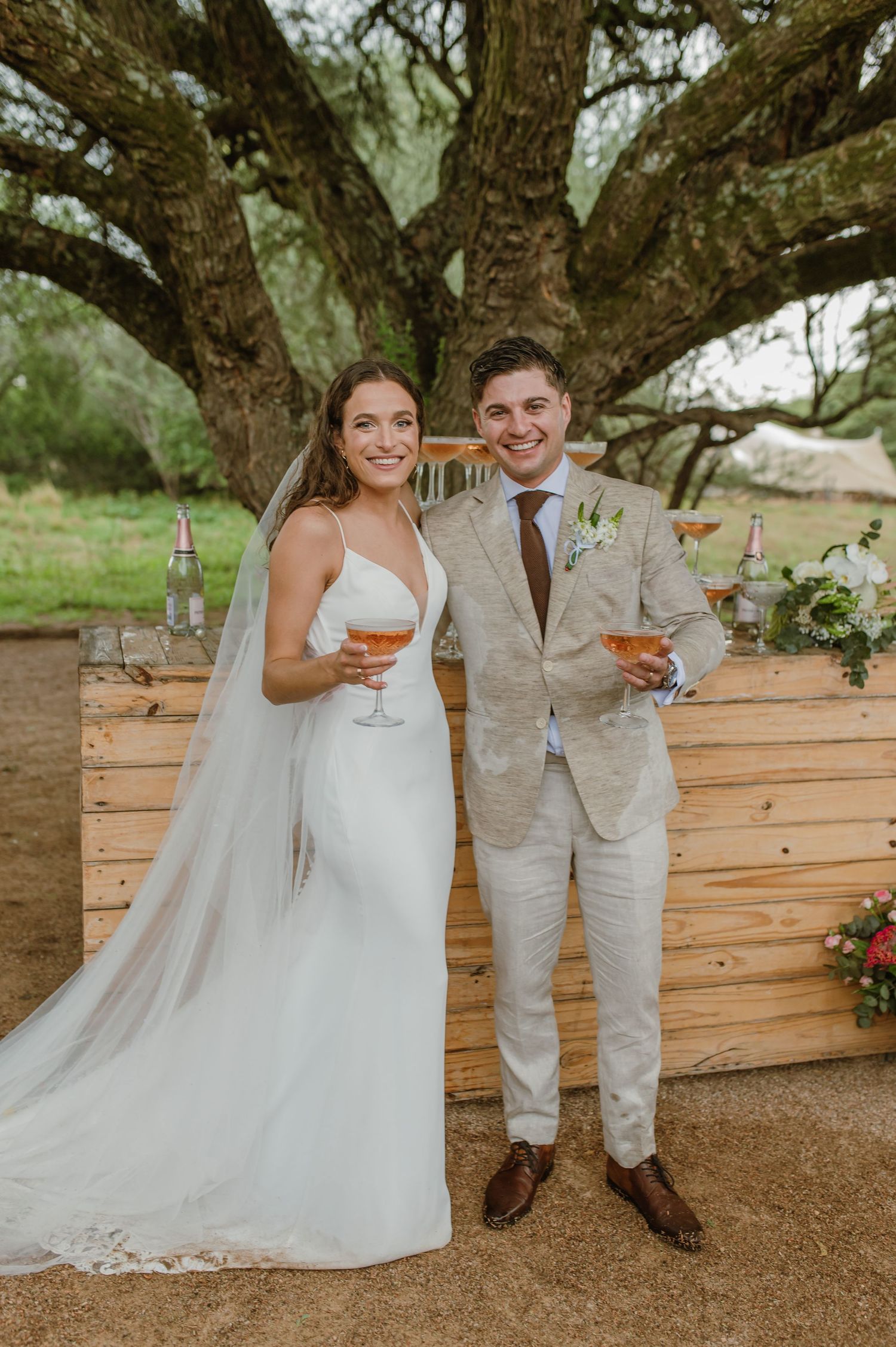 A couple in wedding attire shares a joyful moment under a large tree during an outdoor celebration.