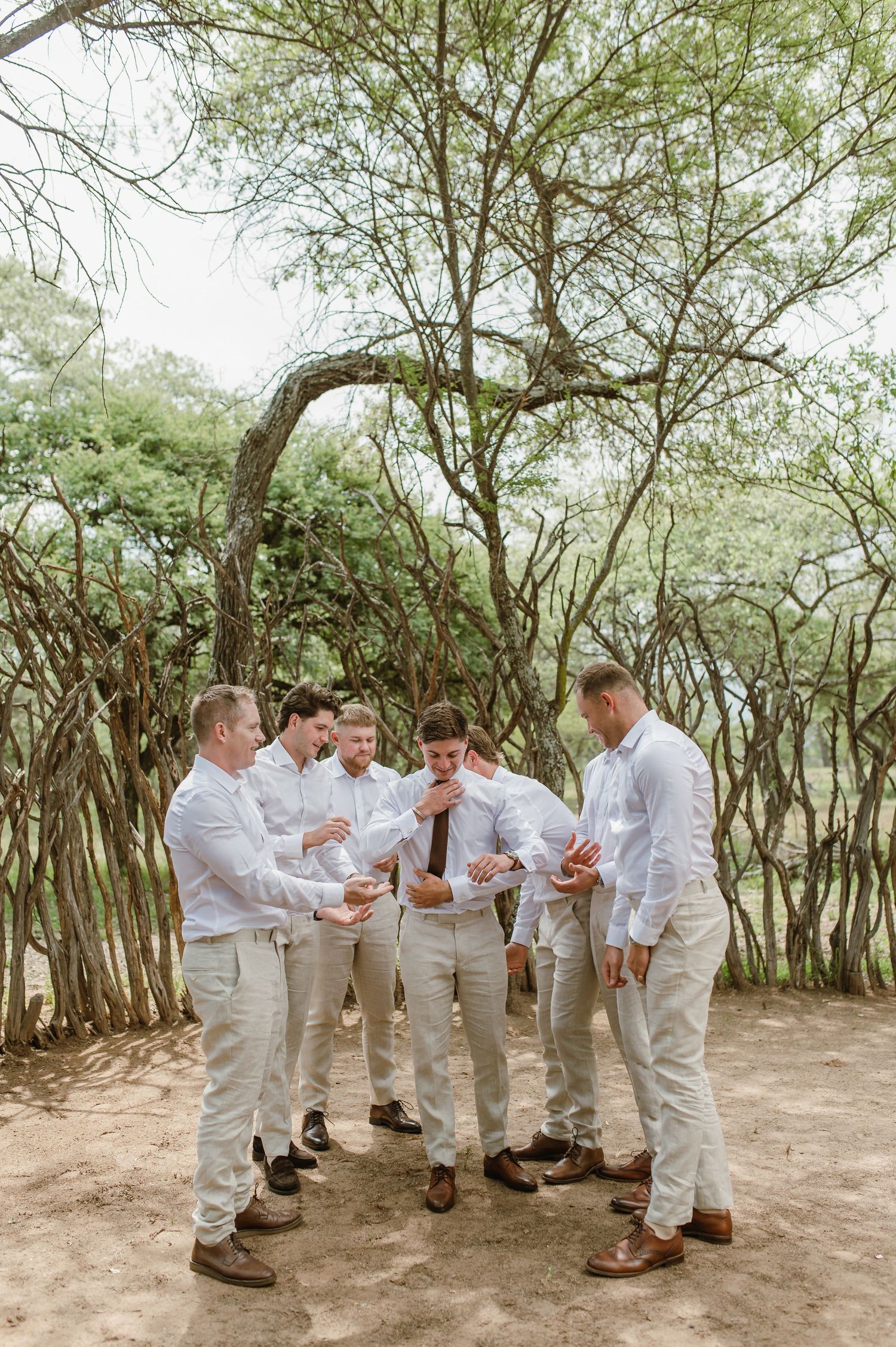 Groomsmen in white suits gather in a circle formation among trees.