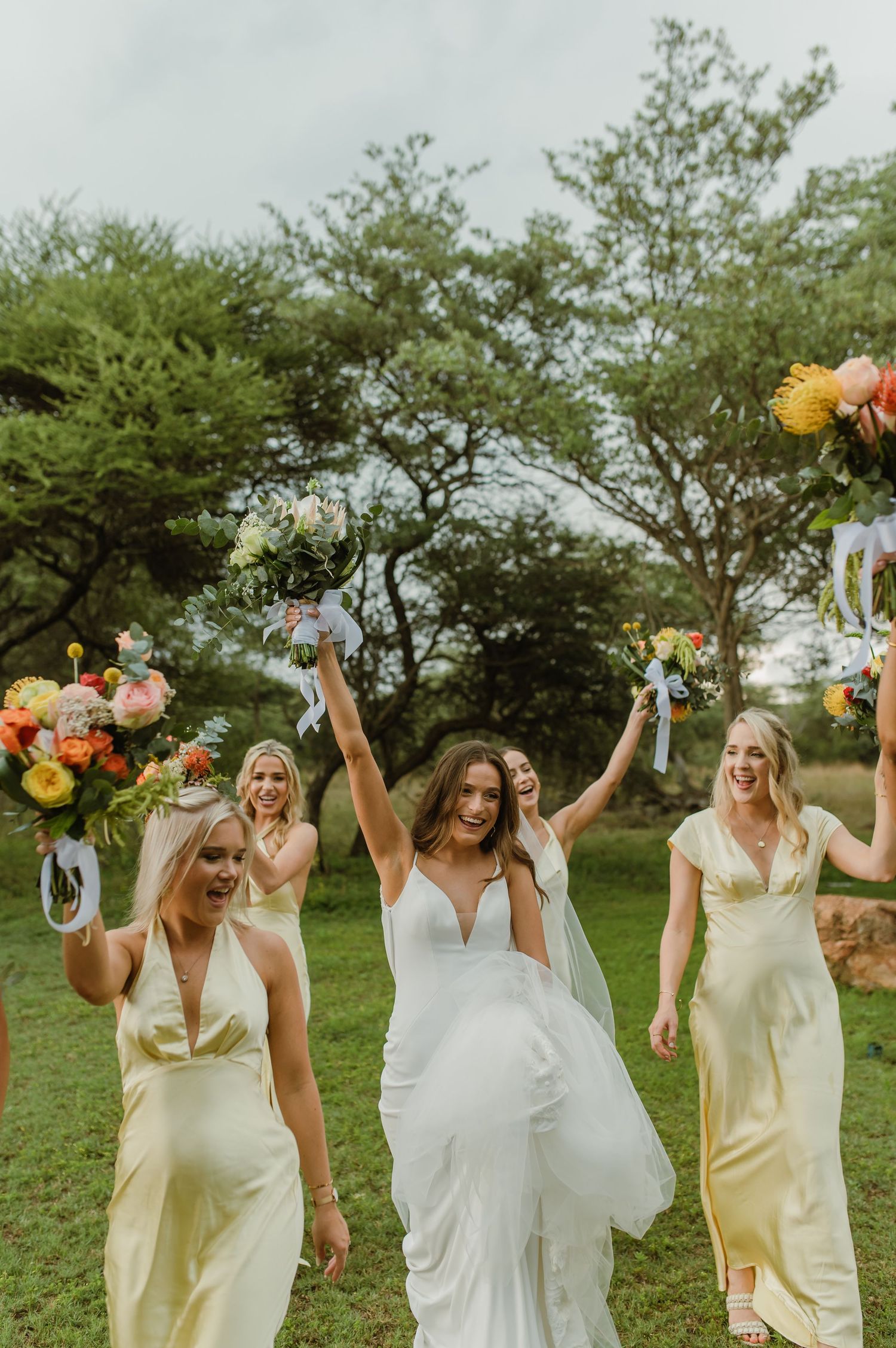 Bridal party celebrates joyfully on lawn with raised bouquets and flowing dresses.