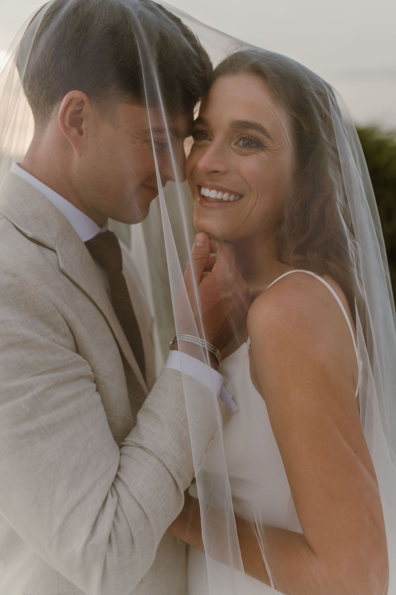 Newlyweds share tender moments beneath a delicate bridal veil during their outdoor photoshoot.