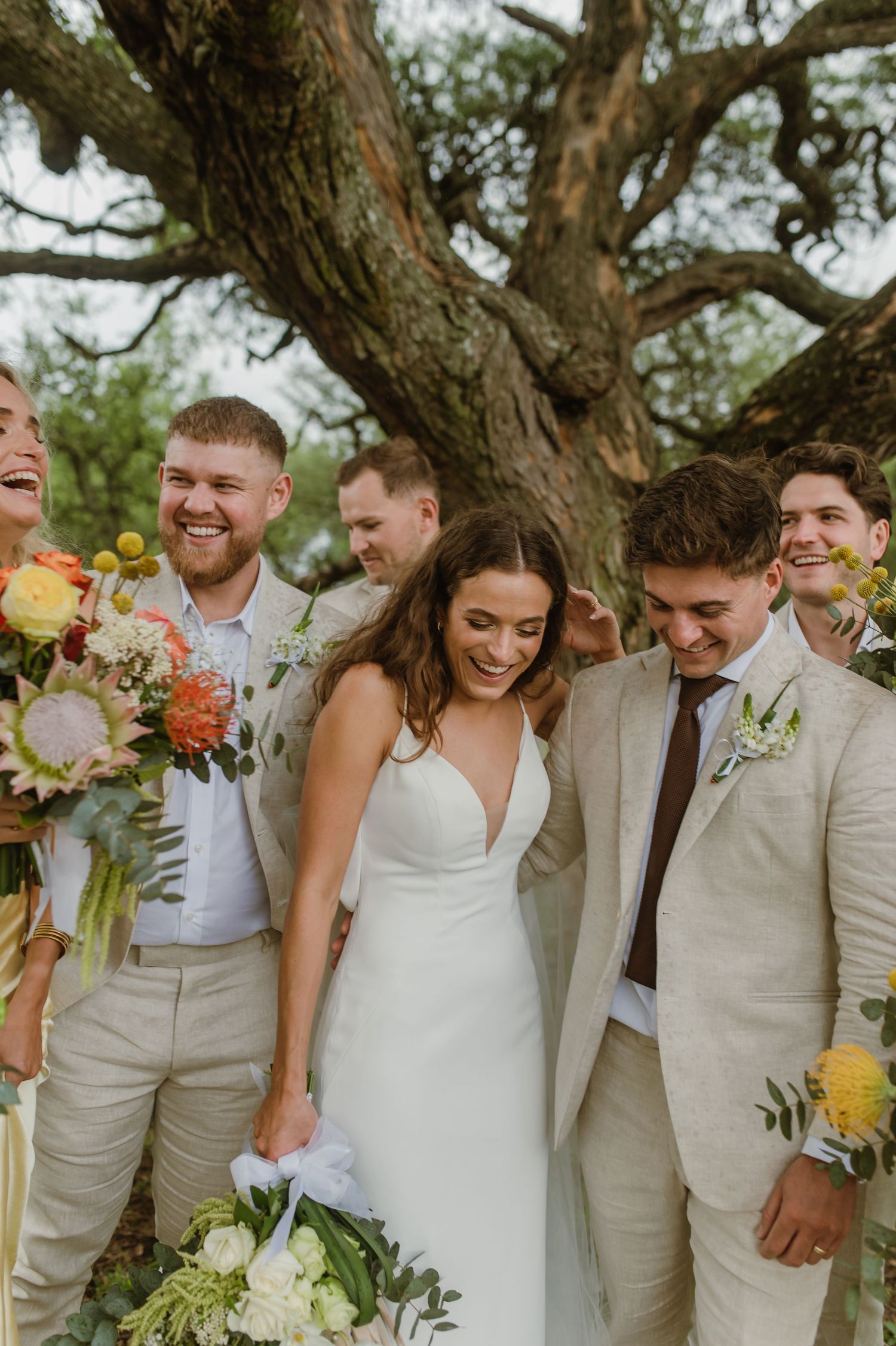 Wedding party shares candid laughs together under a large tree in light-colored attire.