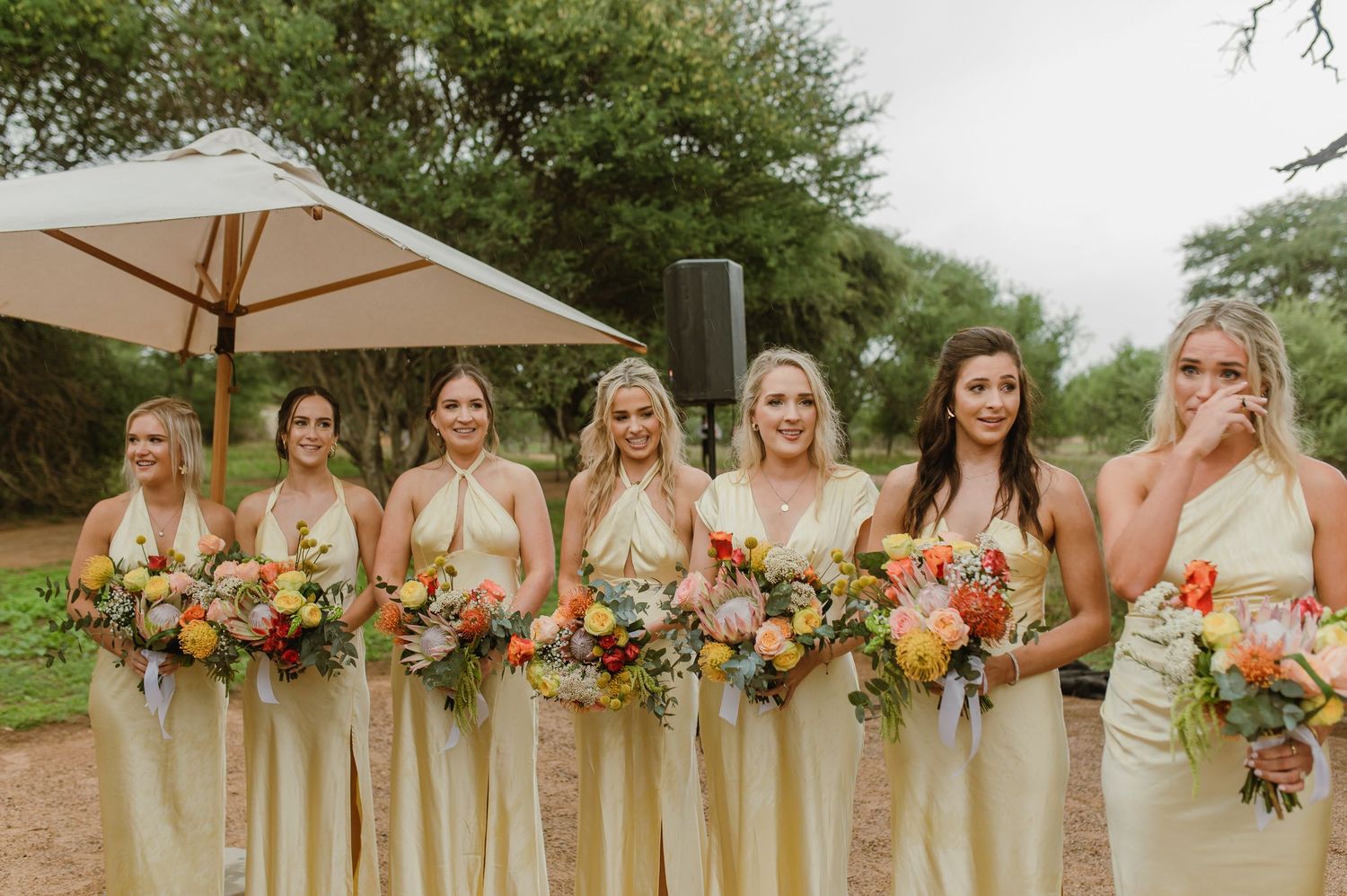 Bridesmaids in neutral dresses hold vibrant orange and pink bouquets at an outdoor wedding.