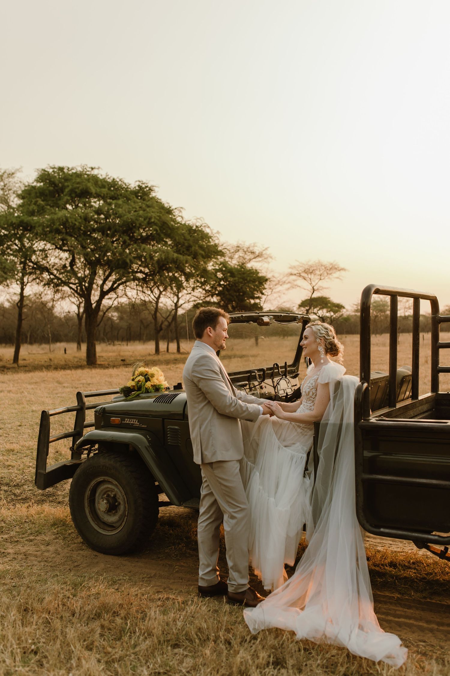 A wedding couple poses beside a vintage safari vehicle in the African wilderness at sunset.