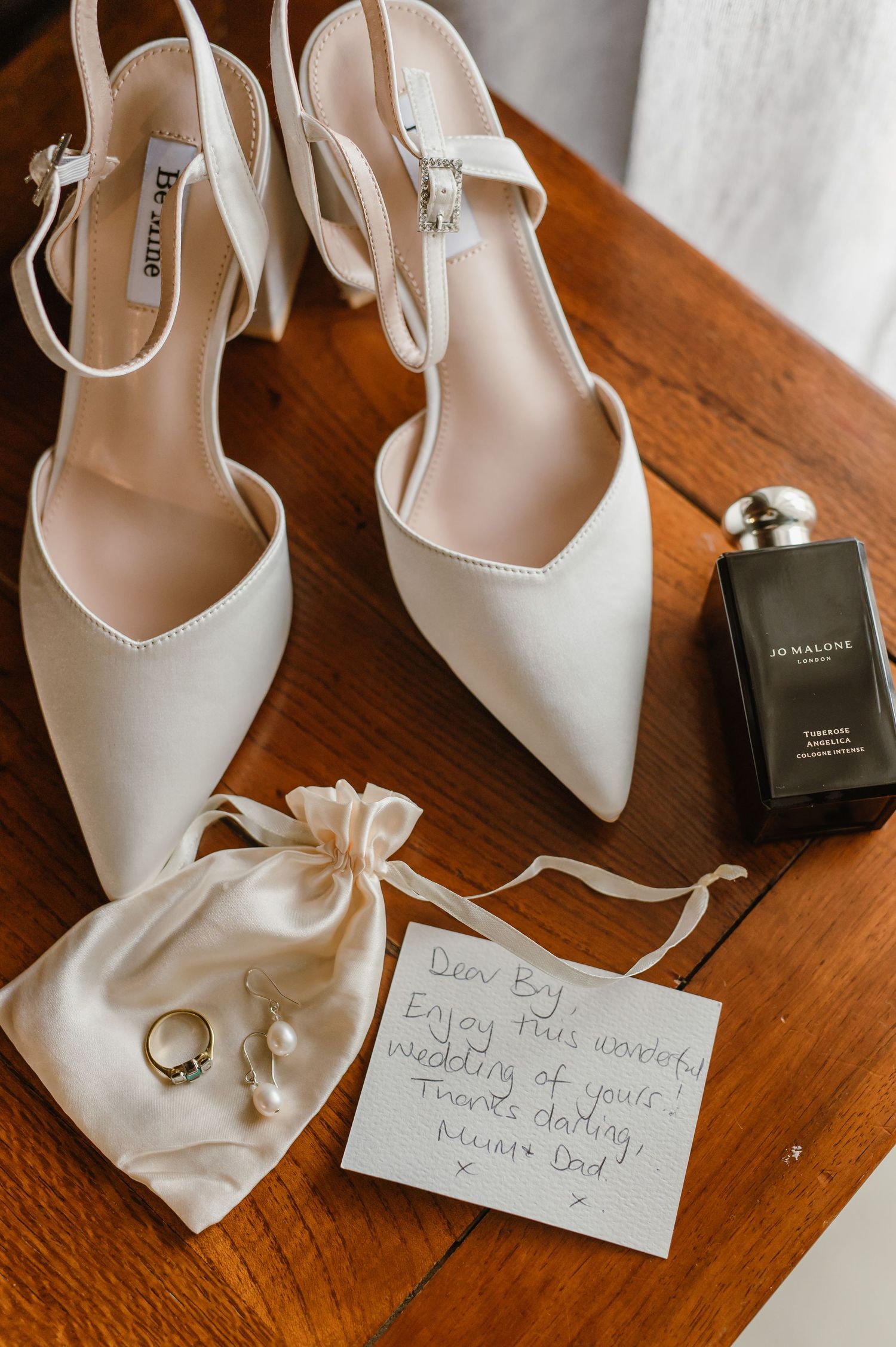 White bridal shoes and handwritten note displayed on wooden surface with jewelry pouch.