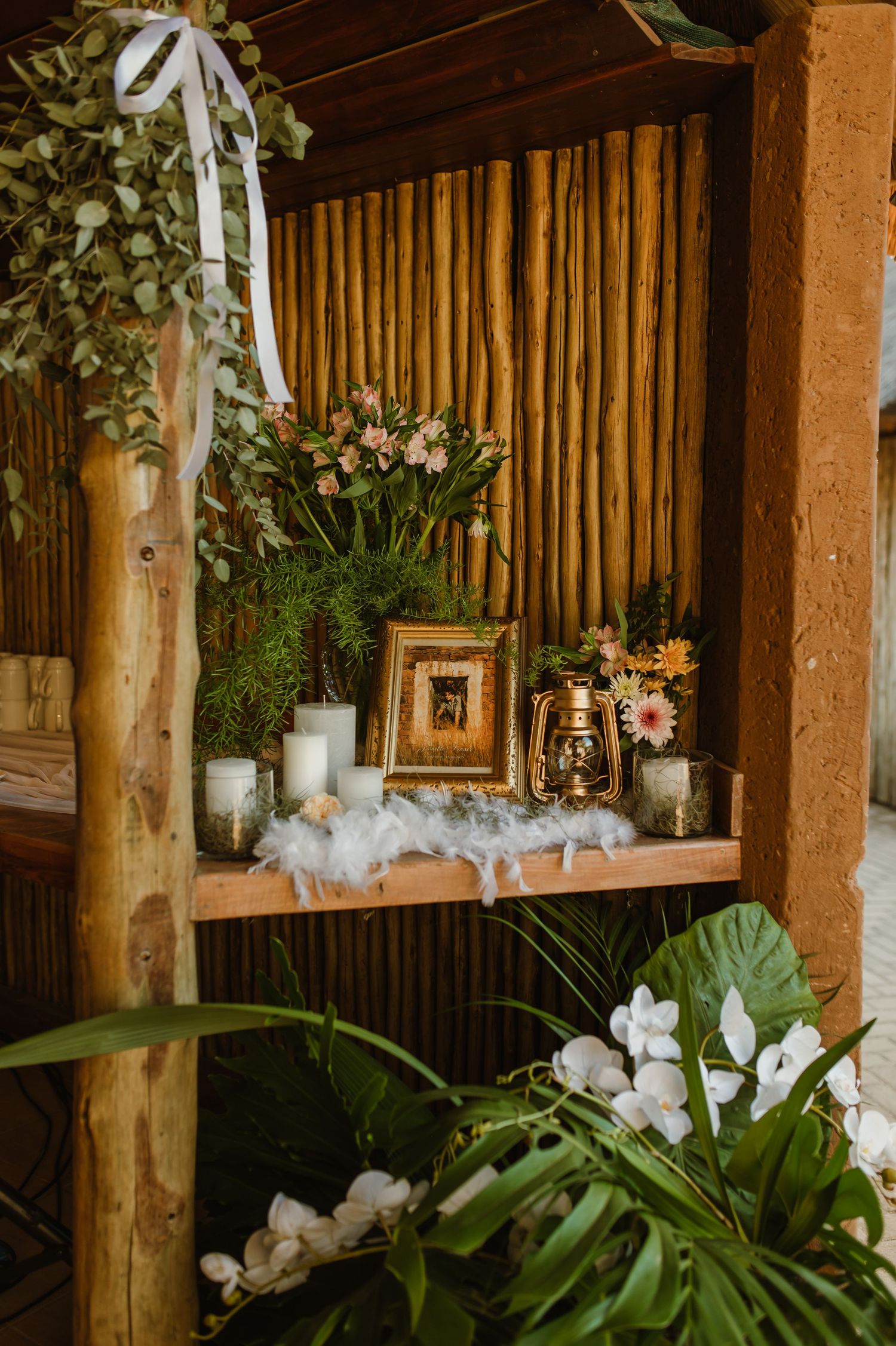 Wedding decor featuring white candles and greenery on wooden shelves in a rustic bamboo setting.