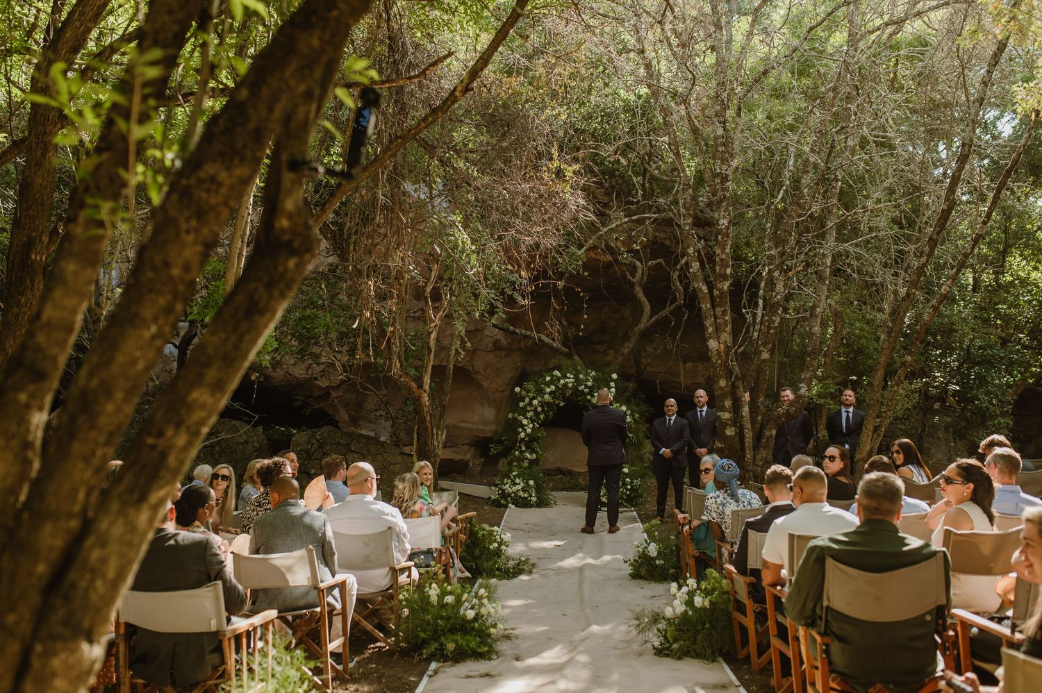 Outdoor wedding ceremony setup with wooden chairs arranged in rows under a canopy of trees.