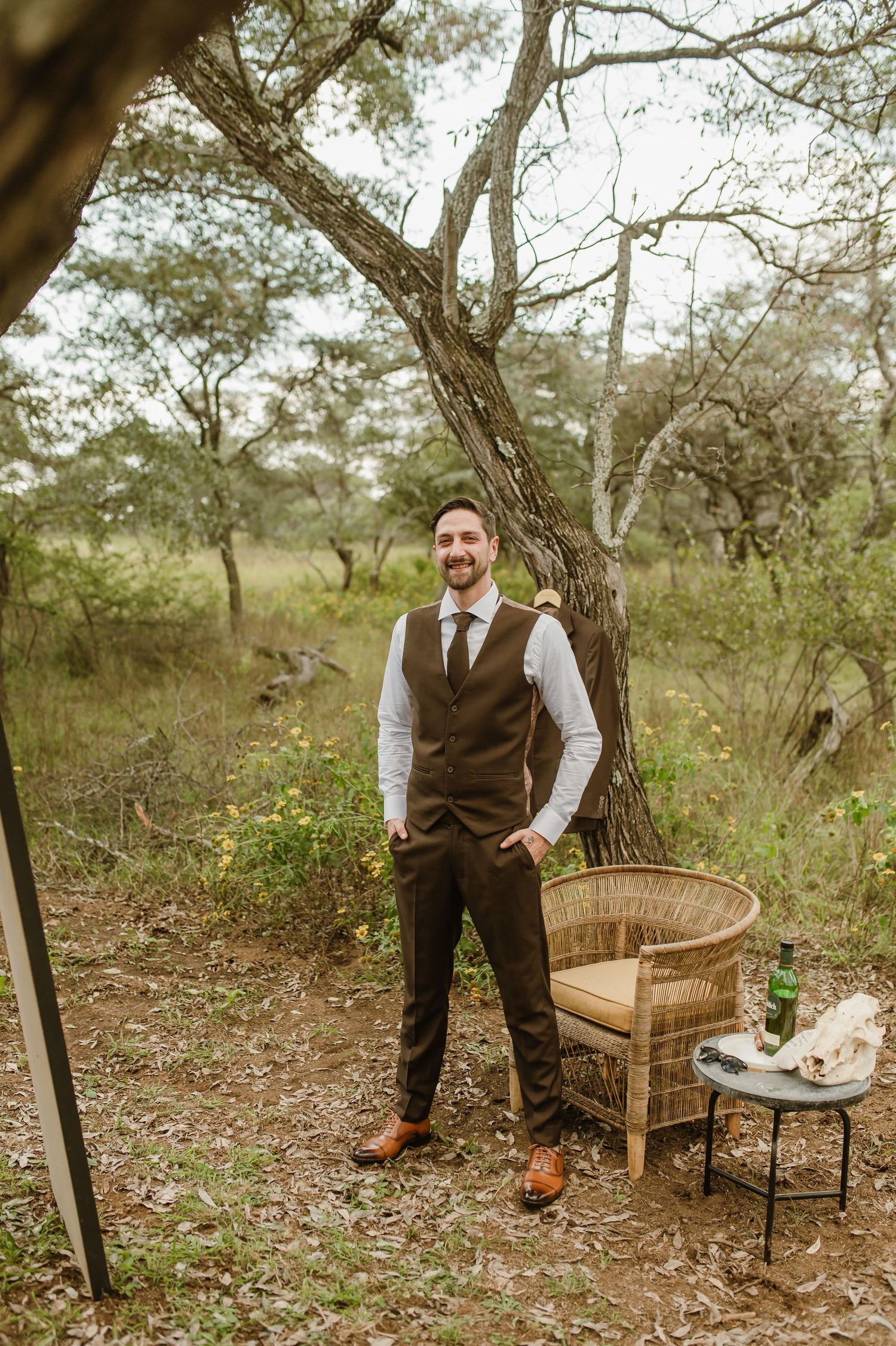 Person in formal attire standing confidently next to wicker chair in natural outdoor setting.