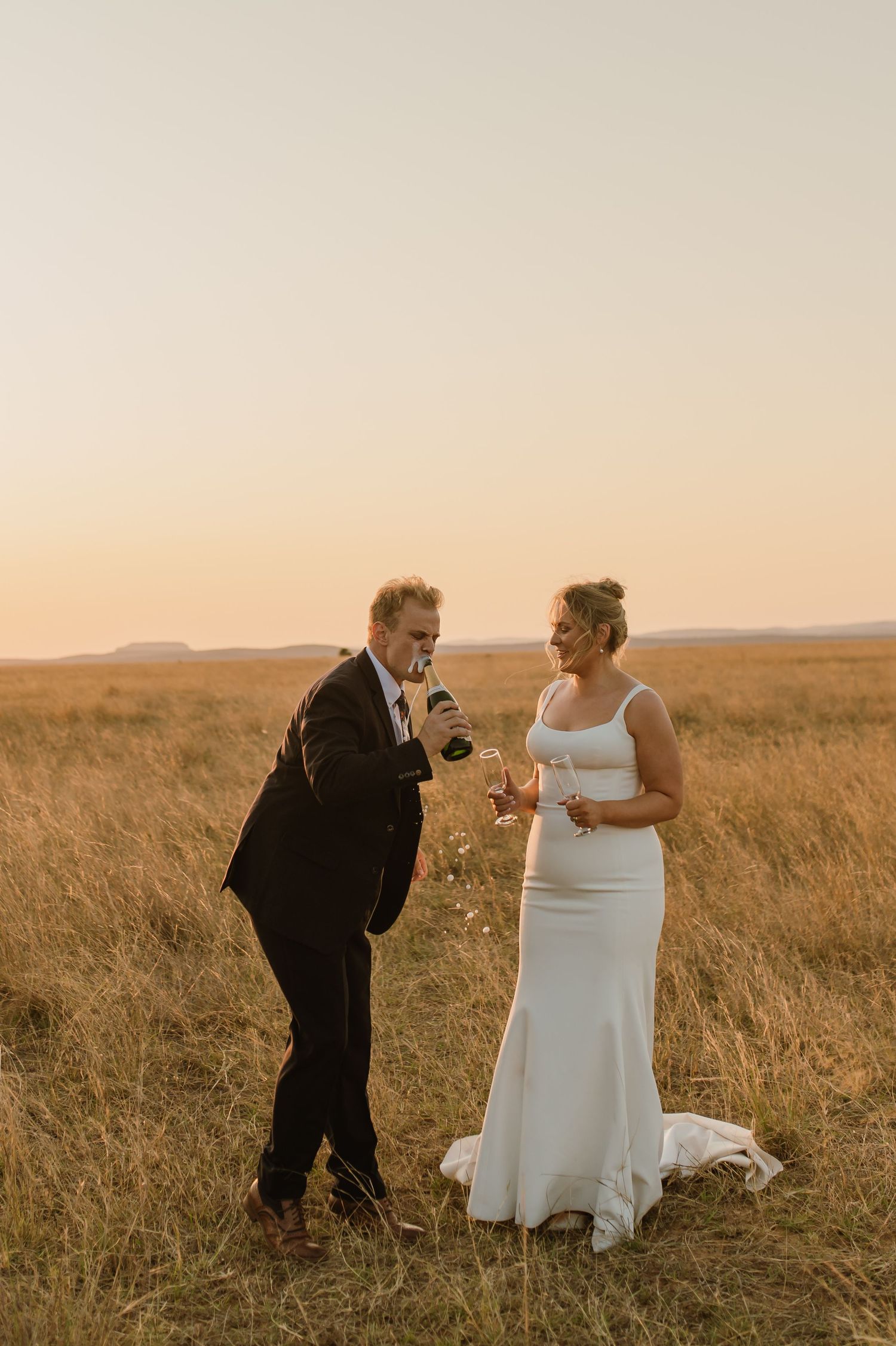 Playful wedding couple interaction in golden grass field at sunset.