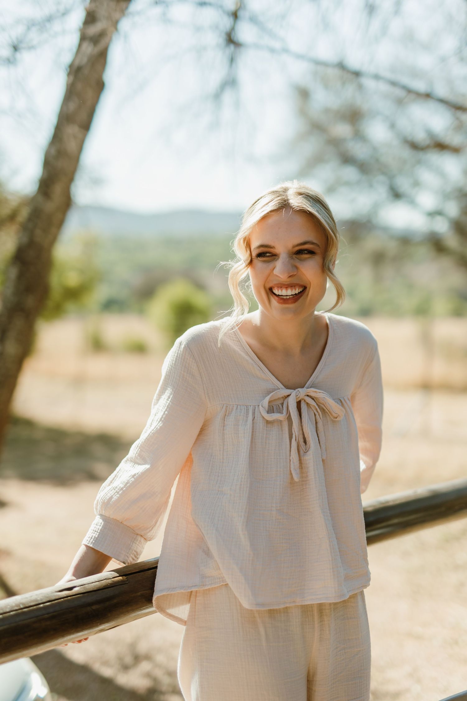 Person in a white flowing dress poses joyfully on a rustic wooden deck in natural outdoor setting.