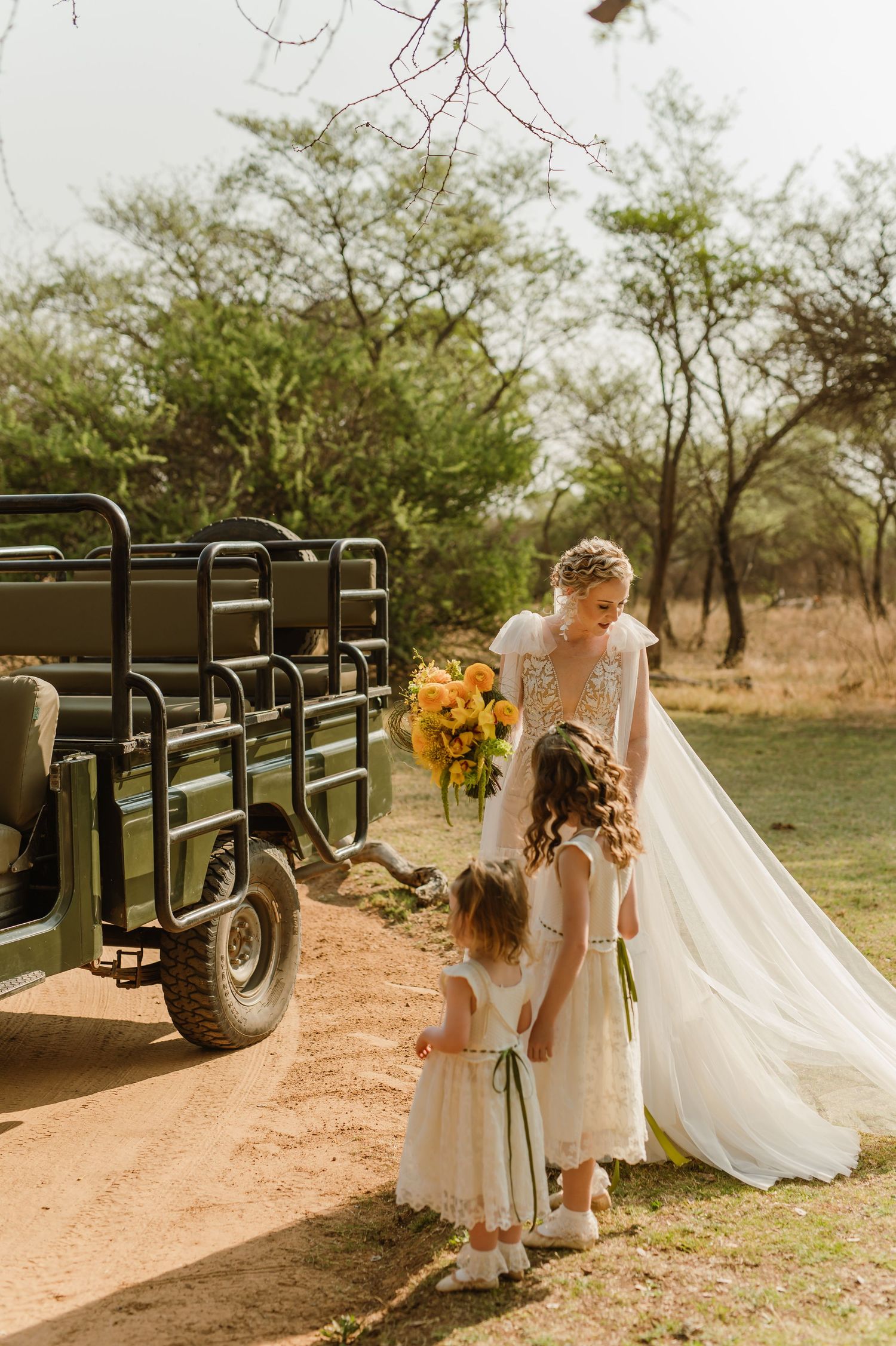 Wedding party poses near safari vehicles on dirt path with flowing bridal veil.