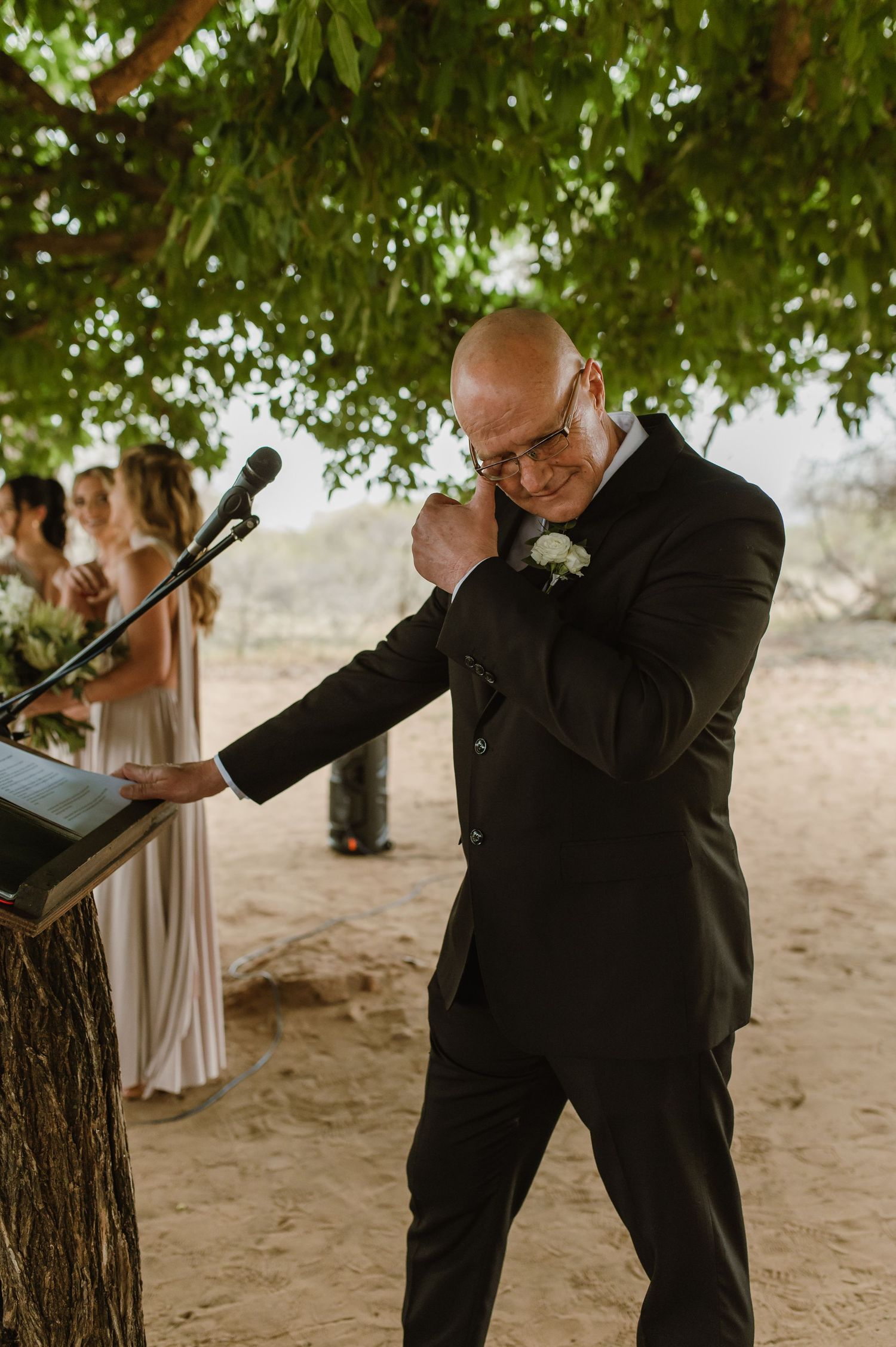 Man in black suit delivering emotional speech during outdoor wedding ceremony.