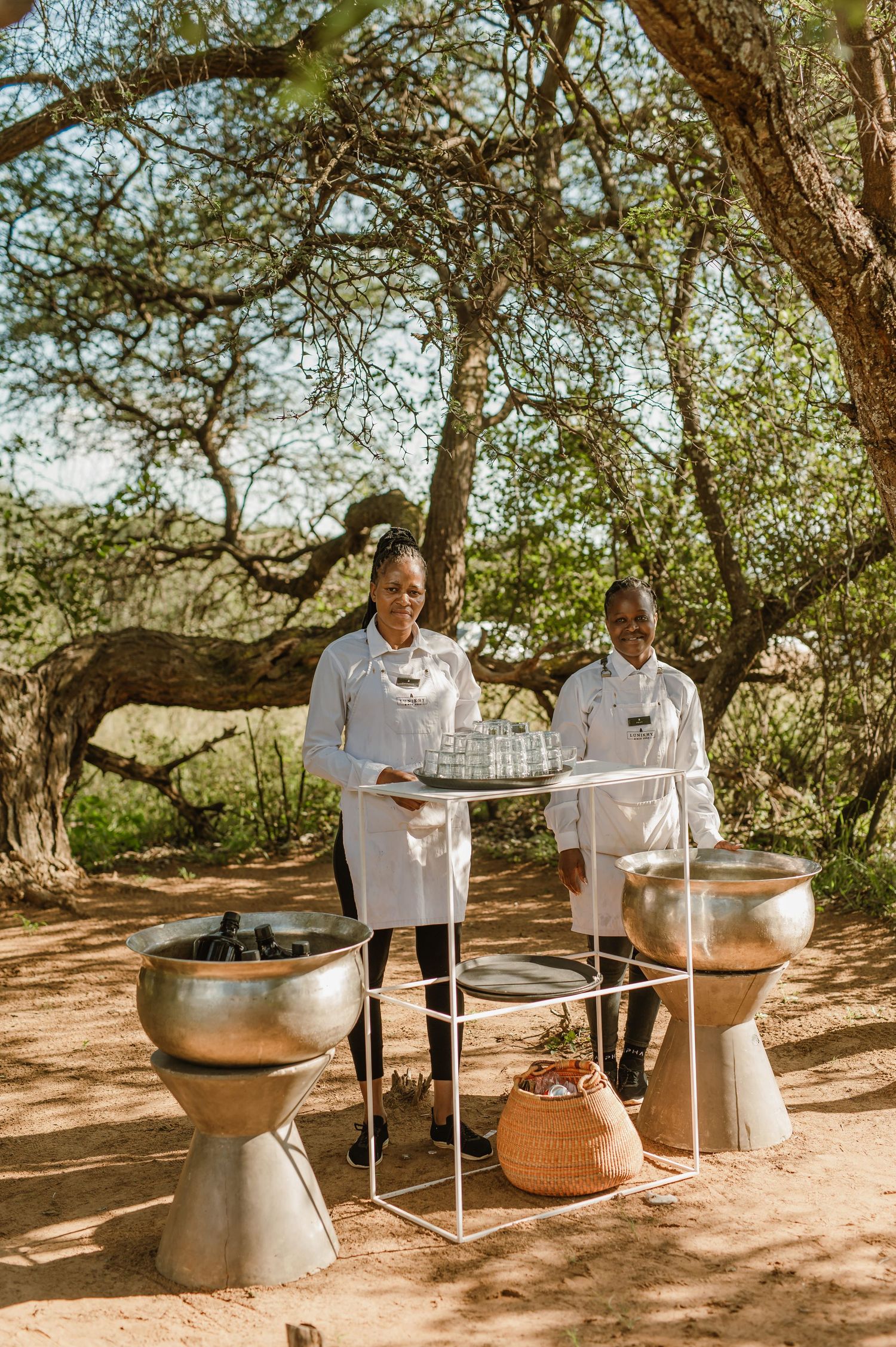 Outdoor cooking station with large pots and chefs preparing food under tree shade.