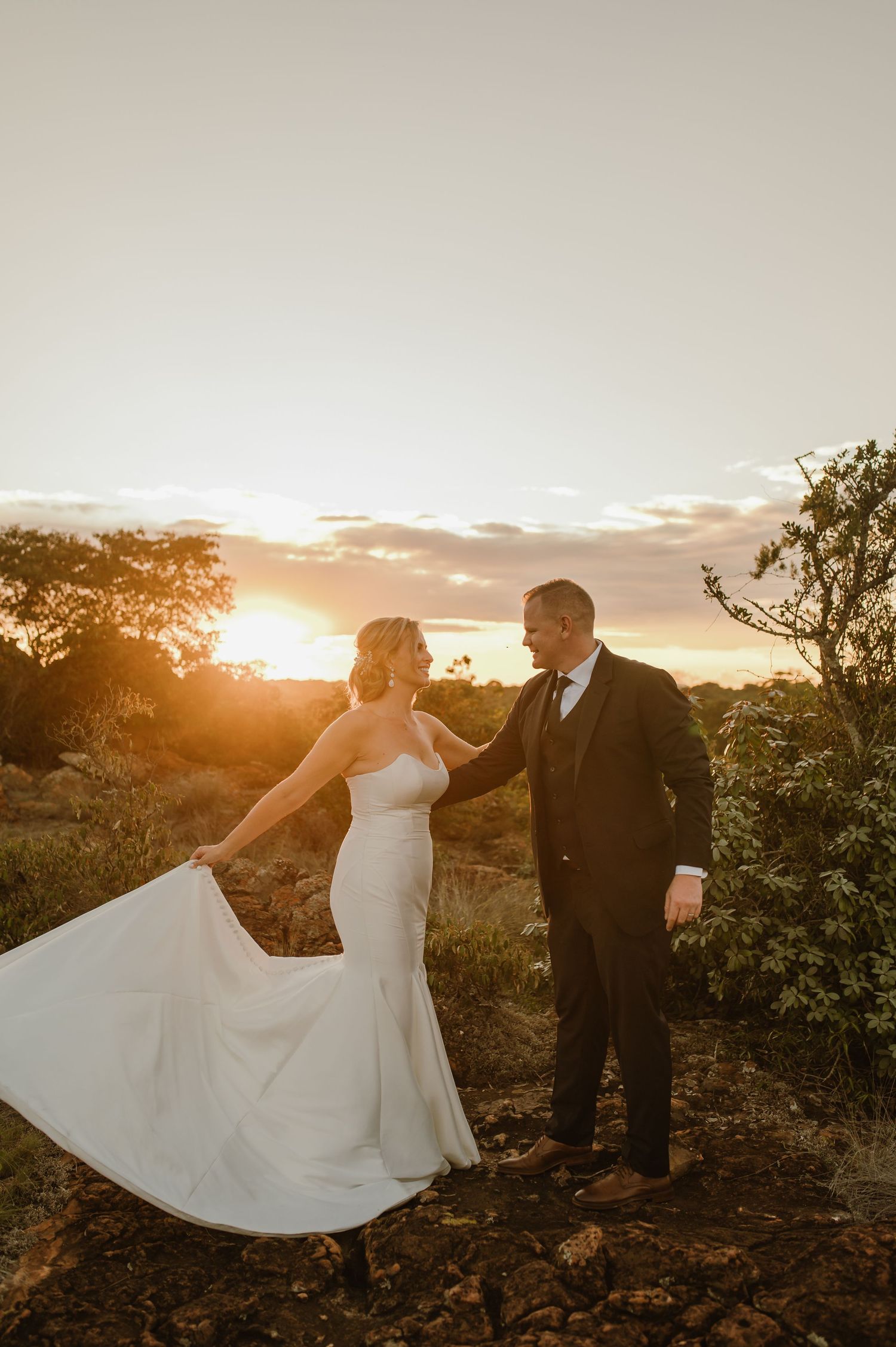 A bride in a flowing white dress twirls with her partner against a sunset backdrop on rocky terrain.
