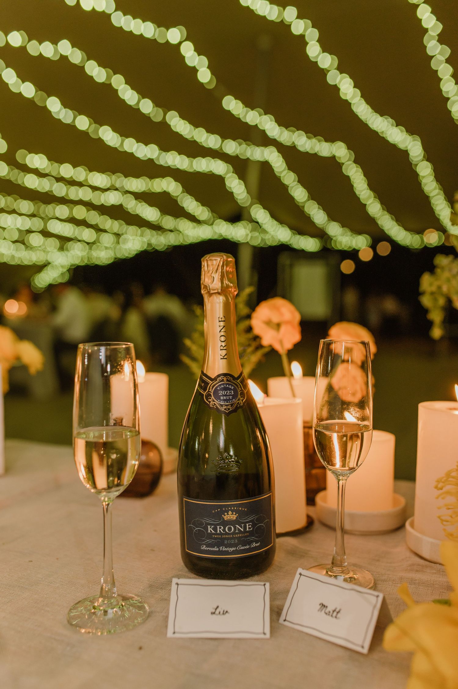 Elegant wedding reception table setting with champagne bottle, glasses and string lights overhead.