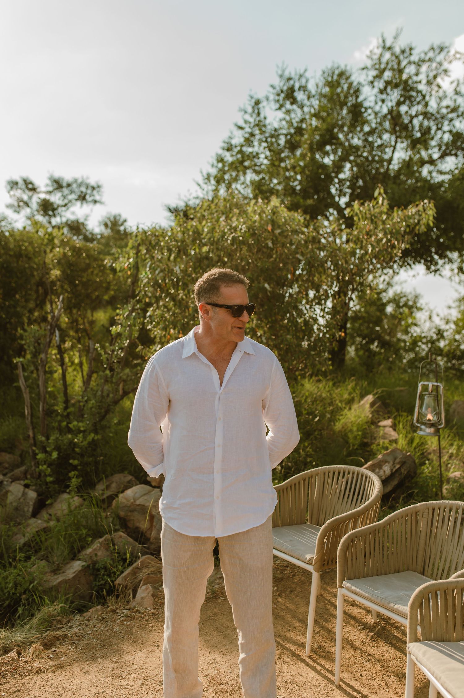 Man in white linen shirt and sunglasses standing outdoors near patio furniture with lush green trees behind him.
