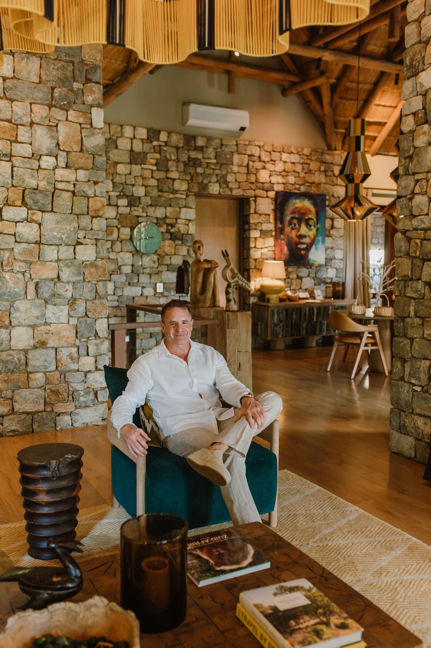 Man relaxing in teal velvet chair in luxury safari lodge with stone walls and African artwork.