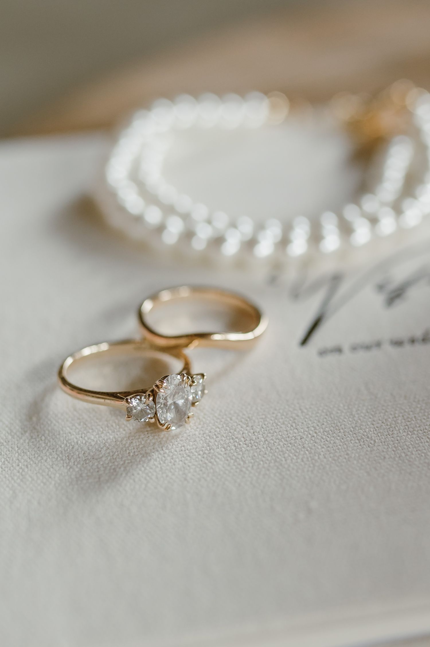 Gold engagement ring with three-stone diamond cluster beside wedding band and pearl necklace.