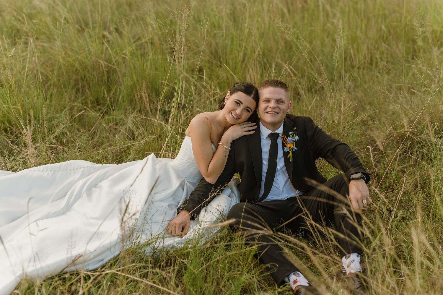 Bride and groom sitting relaxed in tall golden grass field, smiling together in wedding attire.