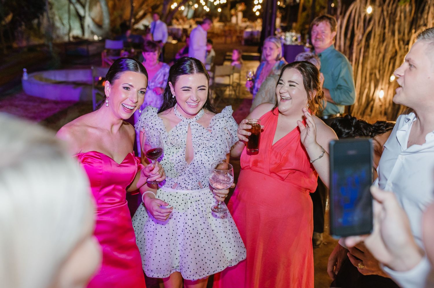 Three wedding guests in pink and white dresses smile and pose together at reception.
