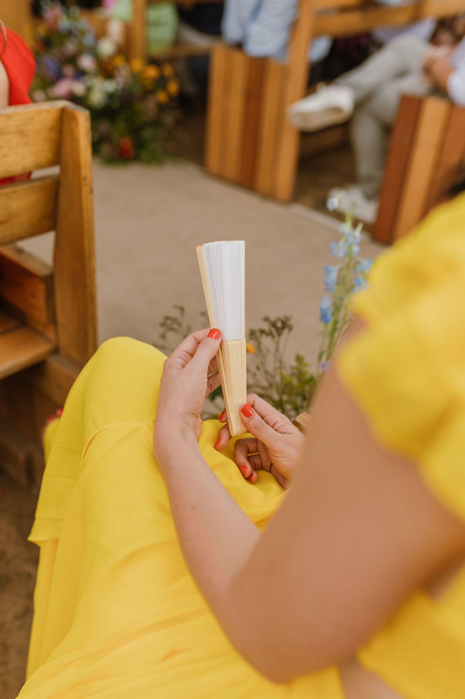 Guest in yellow dress holding a white paper fan at an outdoor wedding ceremony.