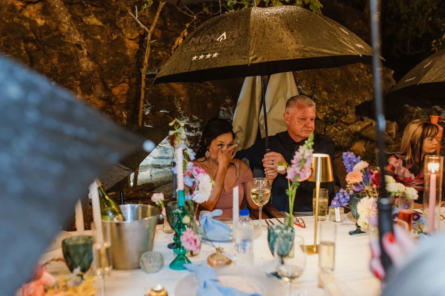 Bride and guest shelter under umbrella at candlelit wedding reception table with colorful floral decor.