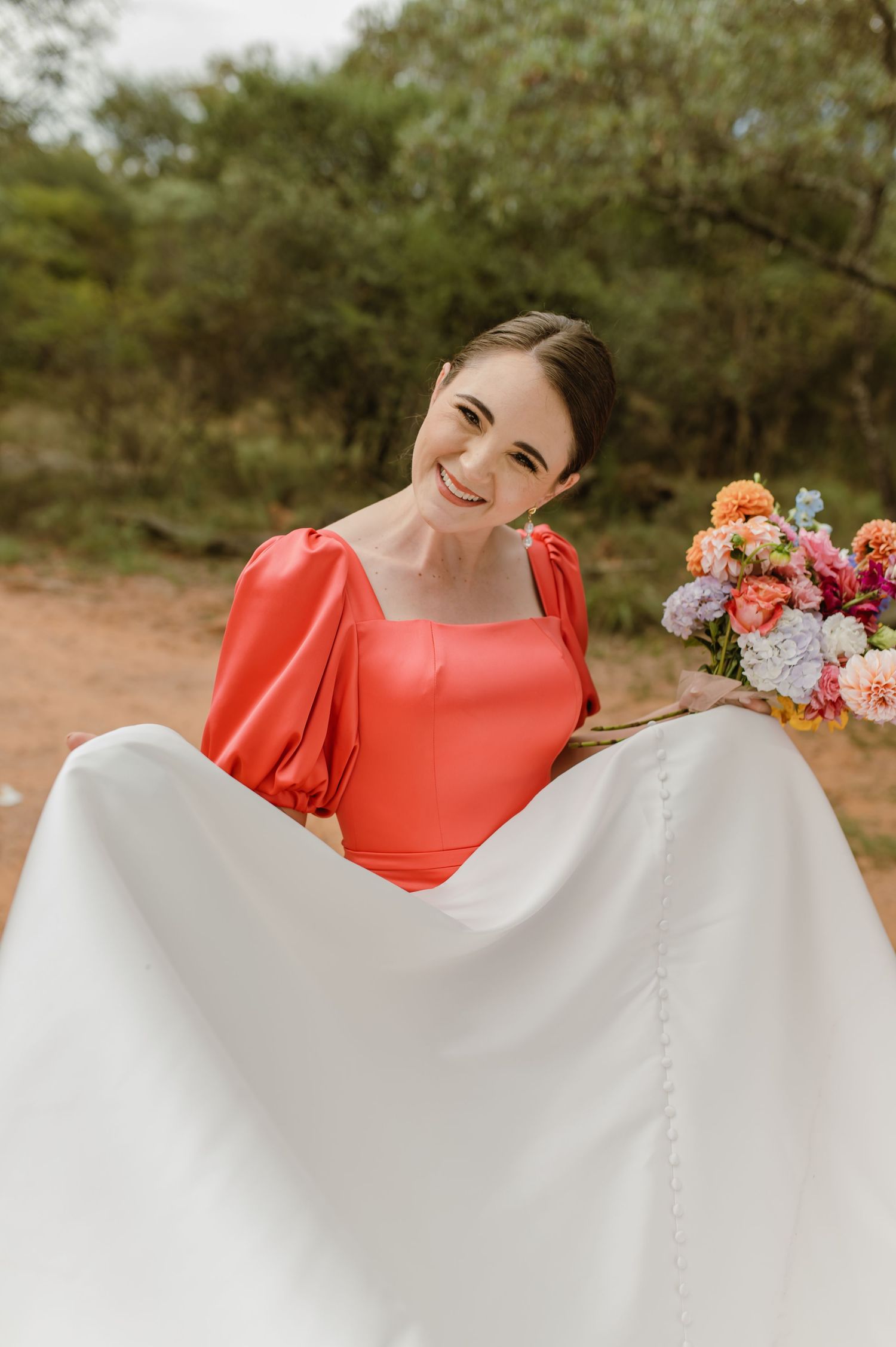 Smiling bridesmaid in red dress holding white wedding gown train and colorful bridal bouquet outdoors.