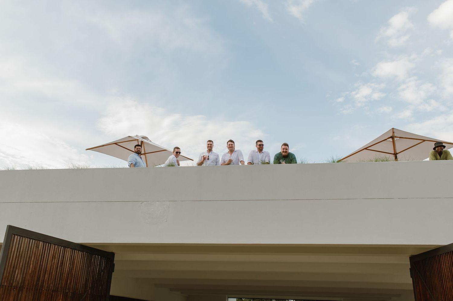 A group stands on a white rooftop under a bright blue sky with beige umbrellas visible in the background.