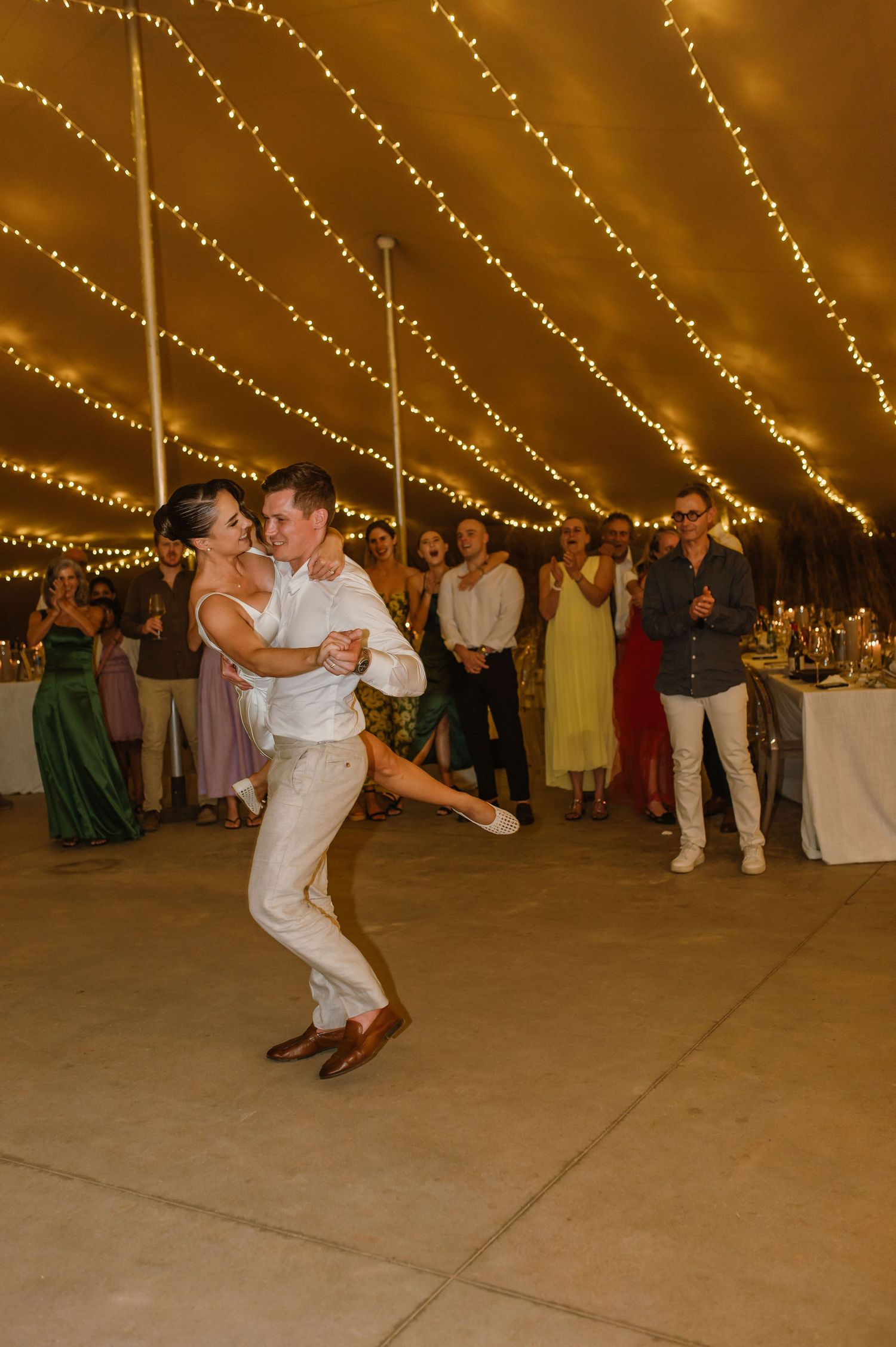 Wedding guests dancing under string lights in a tent with dramatic lighting.