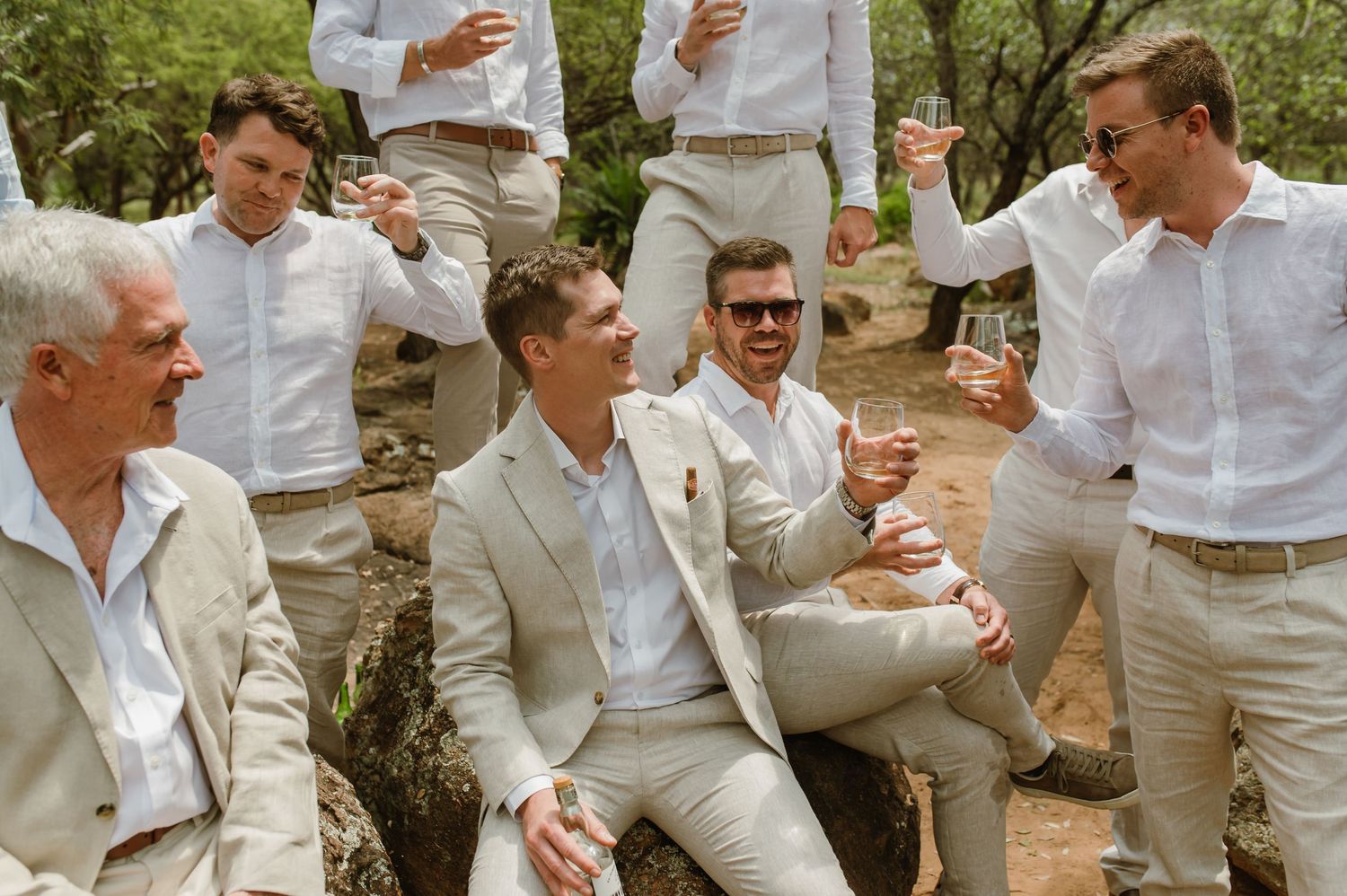 A group in light colored suits and white shirts gather outdoors in a casual celebration setting.