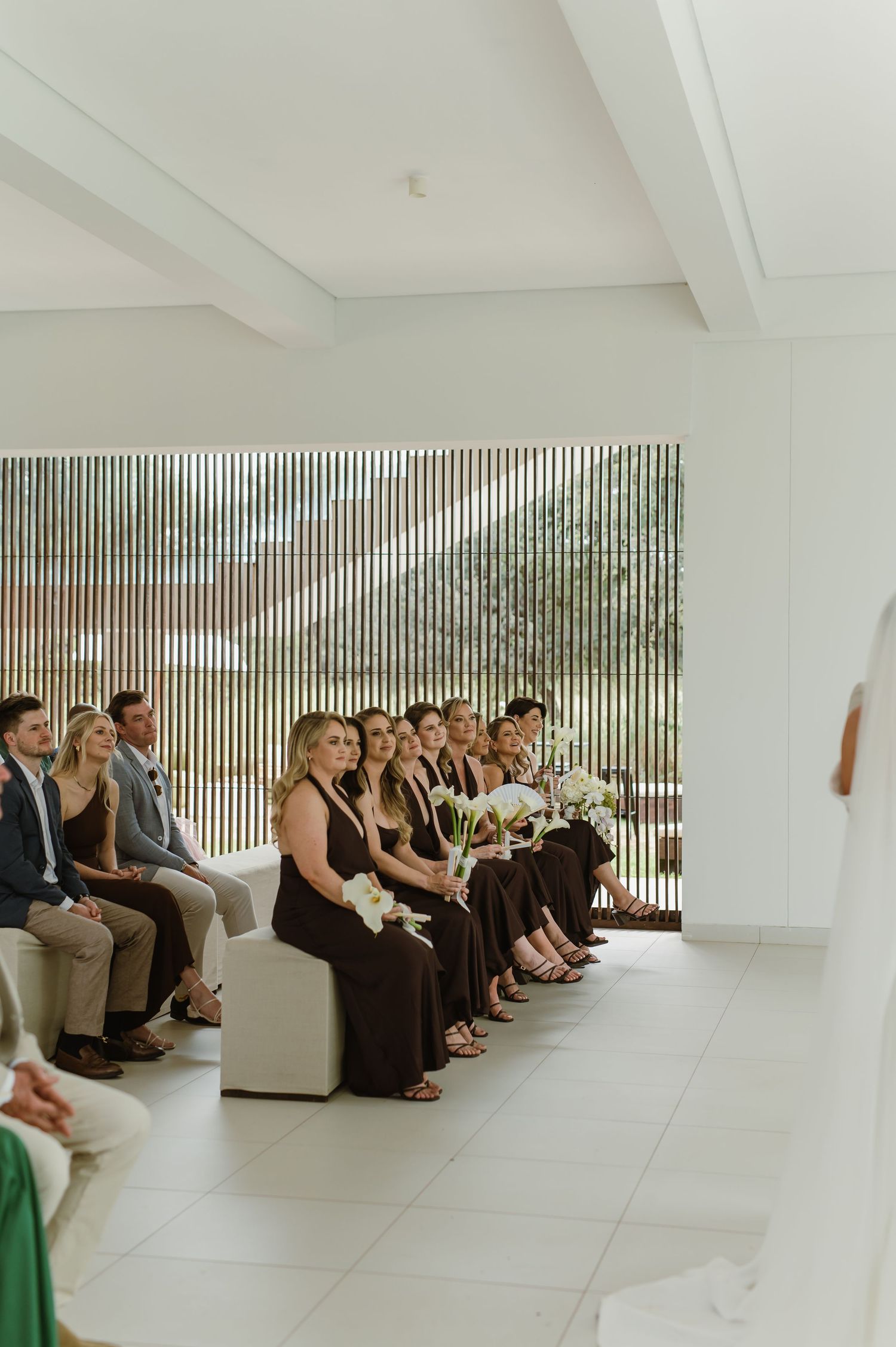 Wedding guests in formal attire seated along a white modern wall with vertical slats.