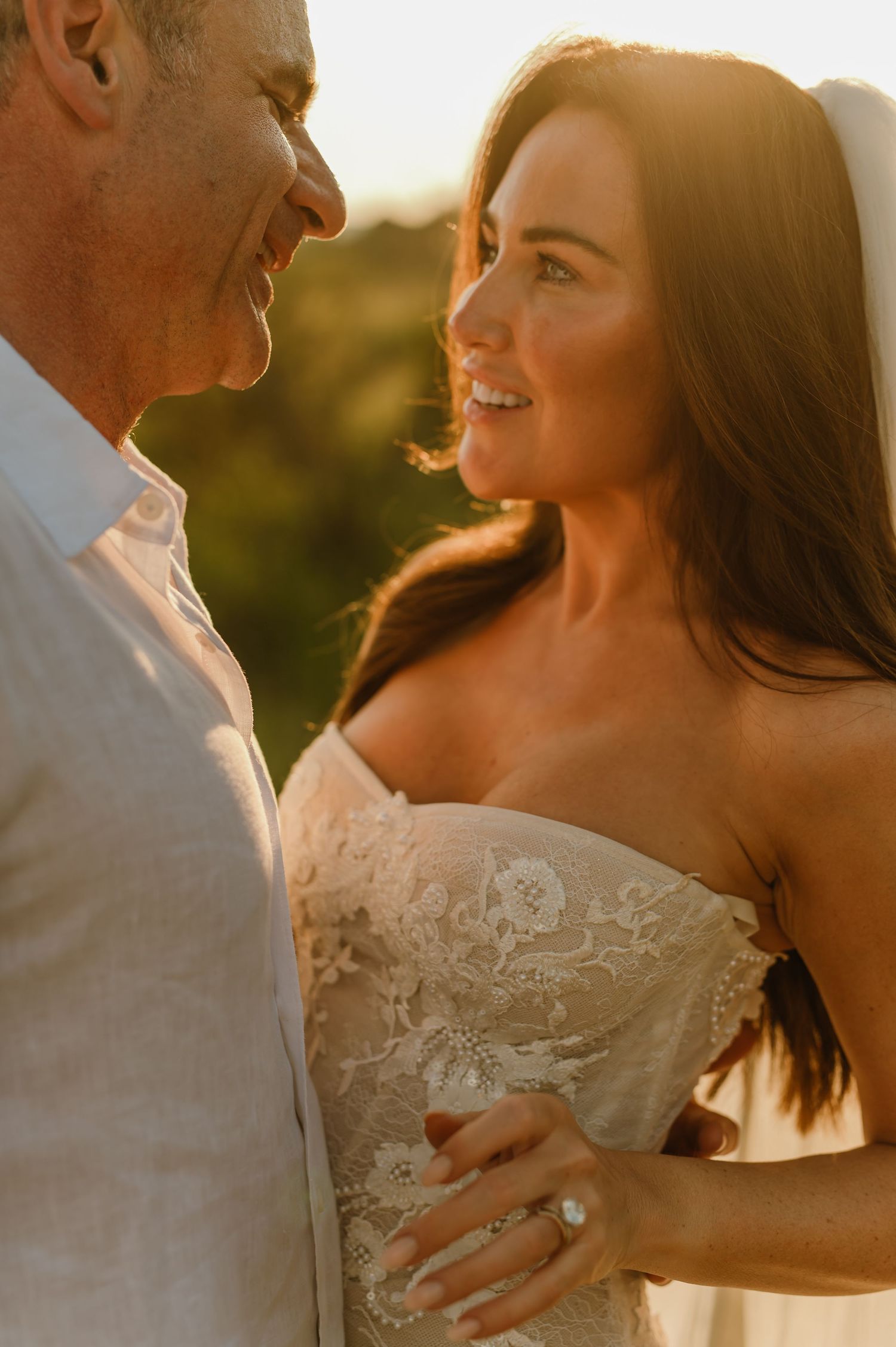 Close-up of smiling bride in strapless lace wedding dress gazing at groom in warm golden sunset light.