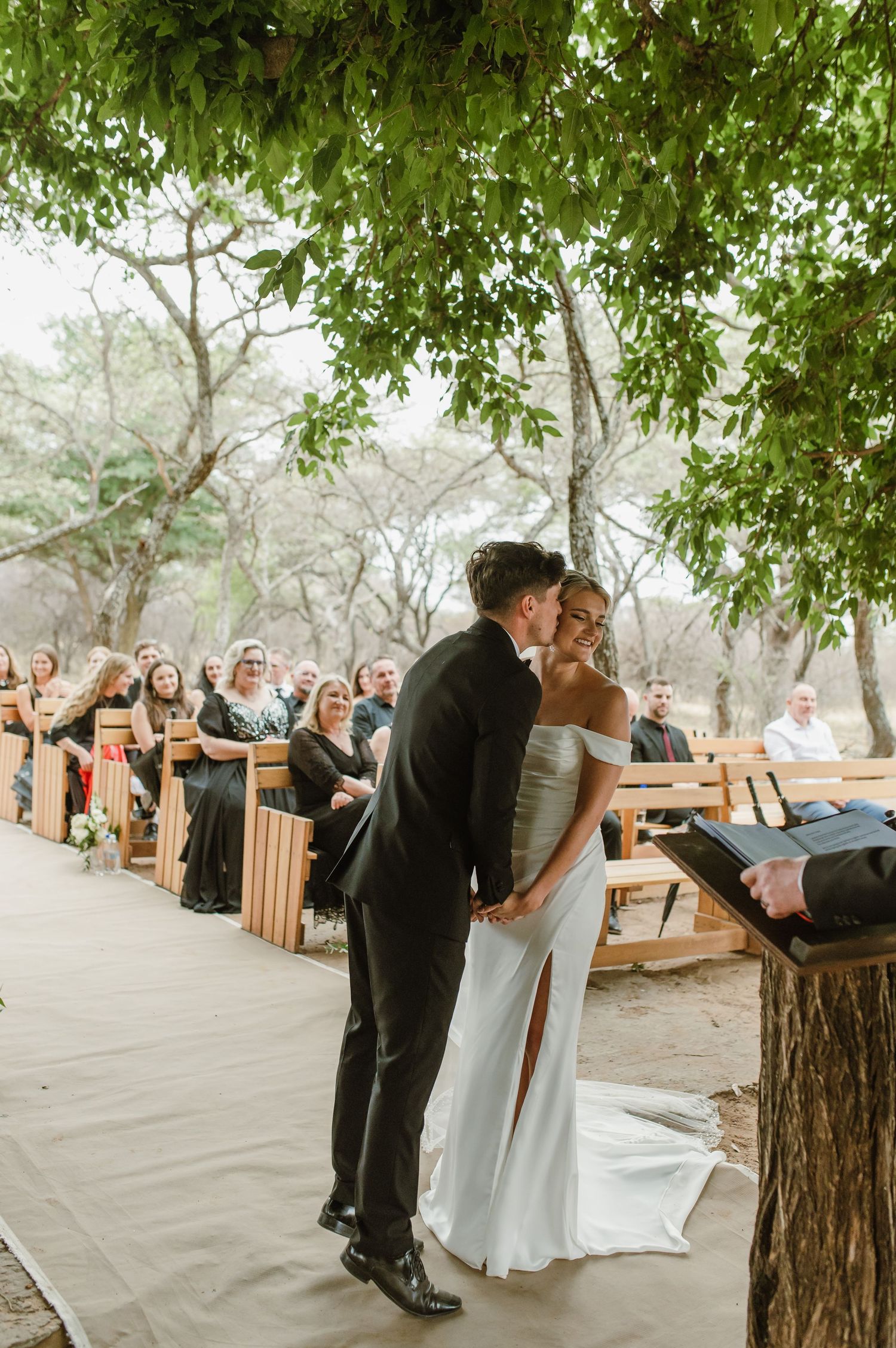 Intimate wedding moment captured in natural outdoor setting with wooden benches and trees.