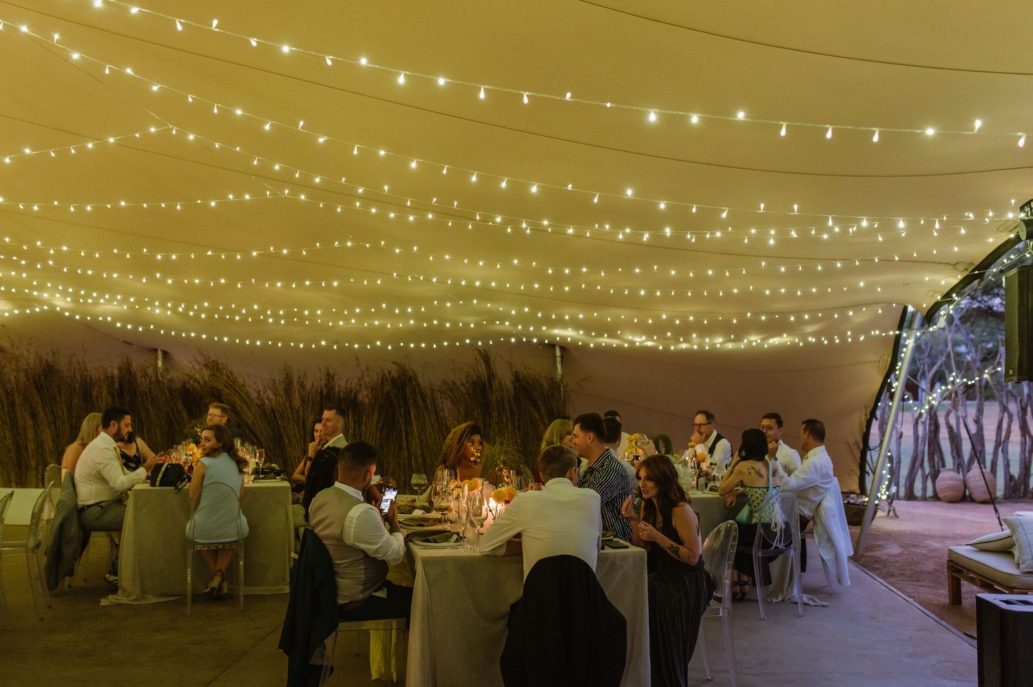 Wedding reception tent decorated with hanging string lights and round tables for guests.