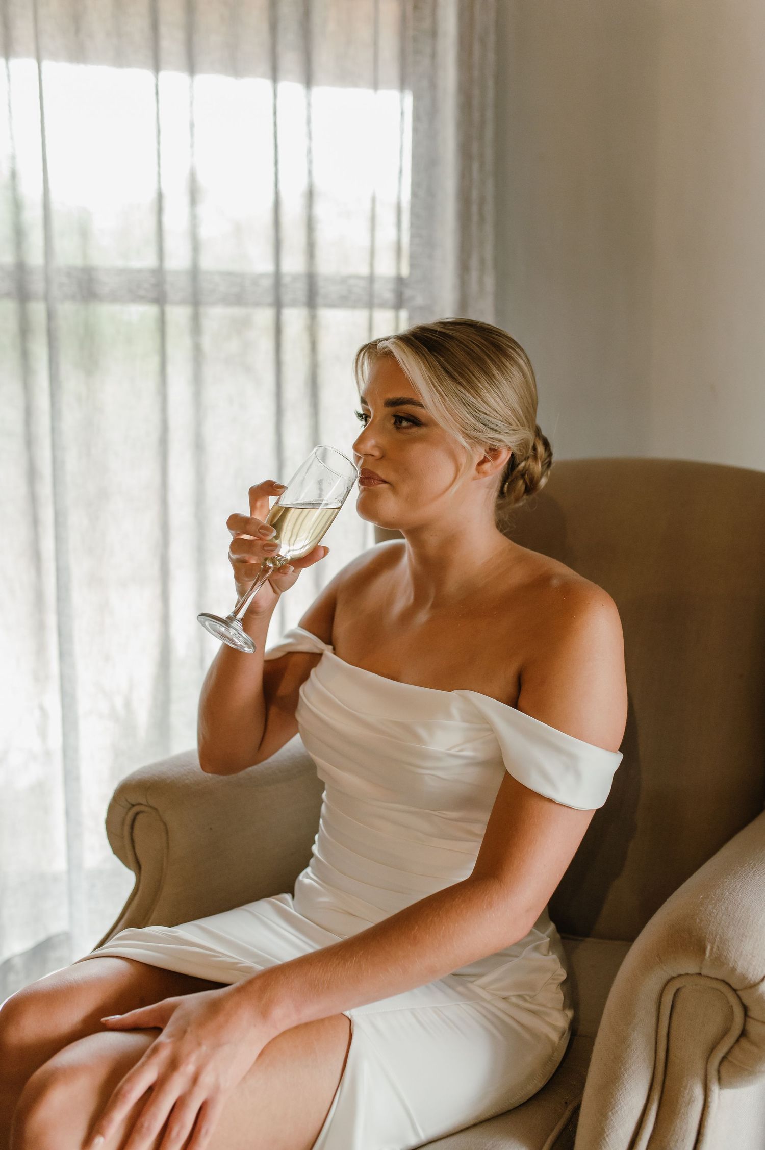 A person in a white off-shoulder dress sits elegantly in a brown armchair holding a glass.