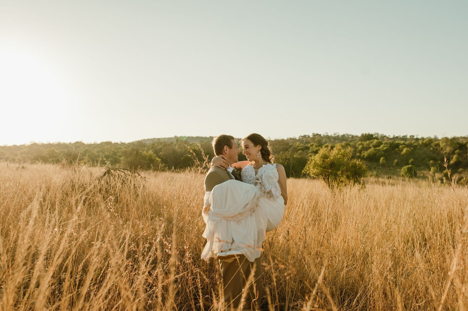 Romantic wedding couple embraces in golden sunlit field during sunset.