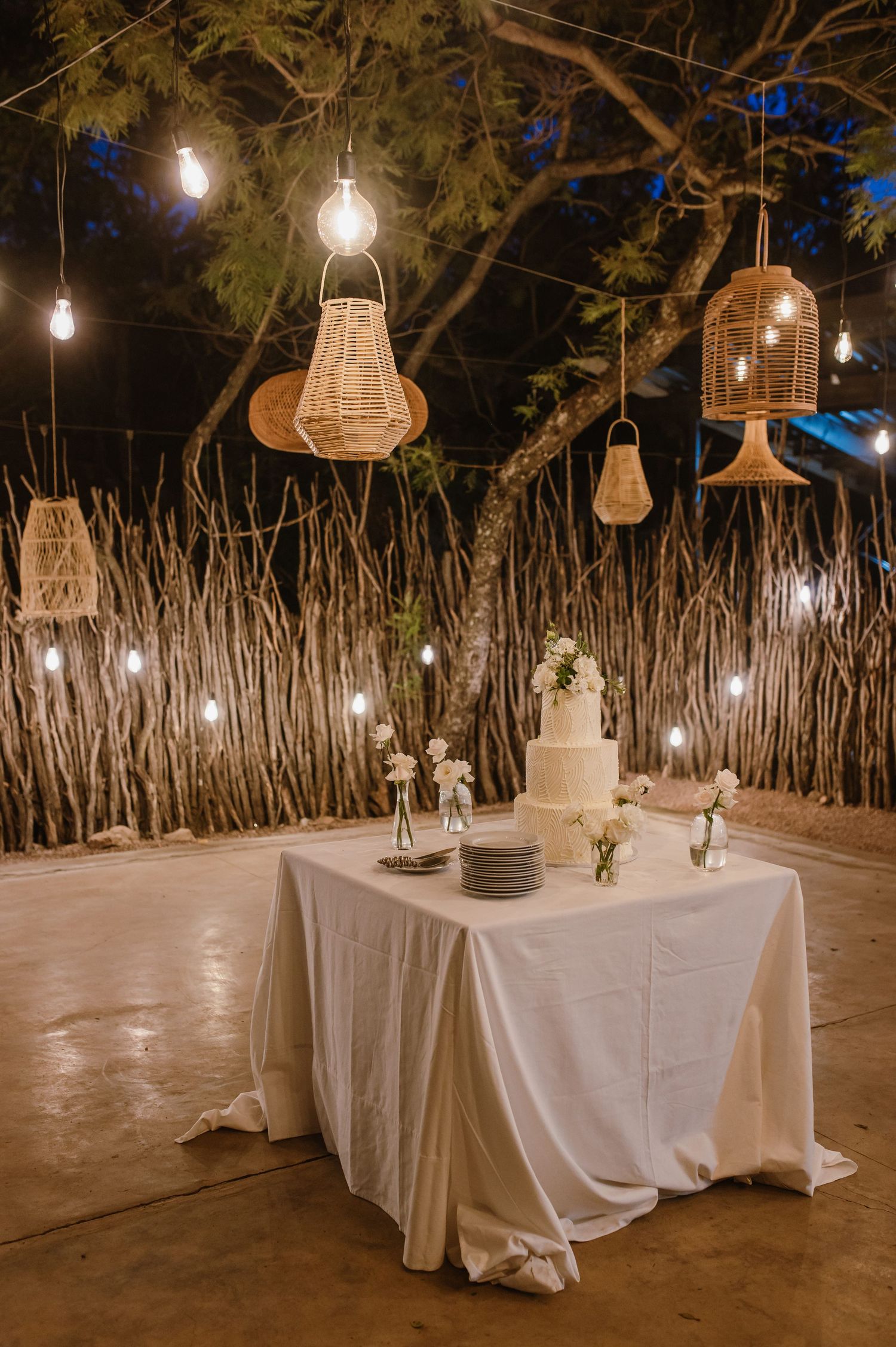 Rustic outdoor dining area with woven pendant lights hanging over white-clothed tables.