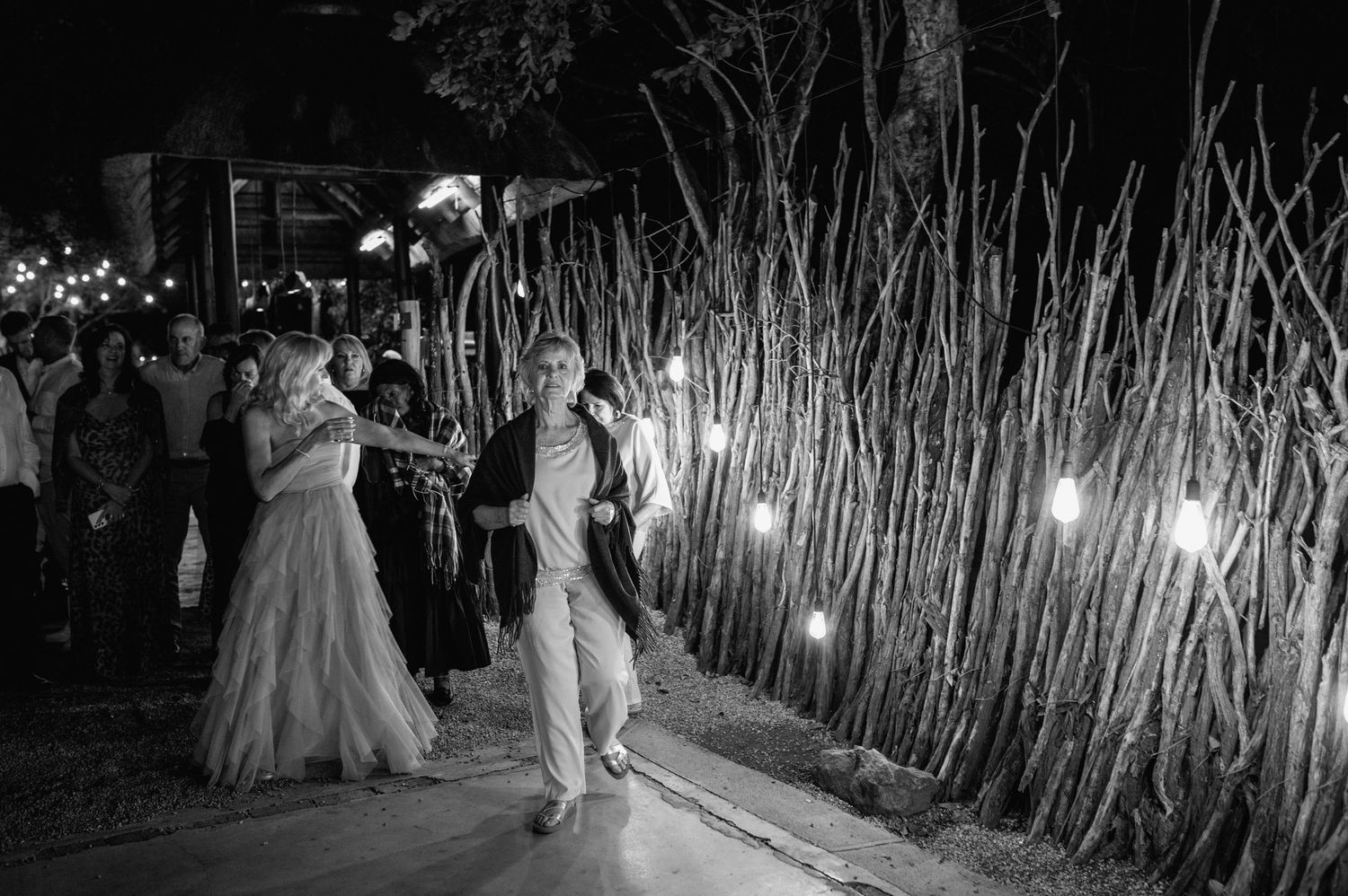 Wedding guests dance and celebrate on an outdoor dance floor illuminated by string lights and surrounded by trees.