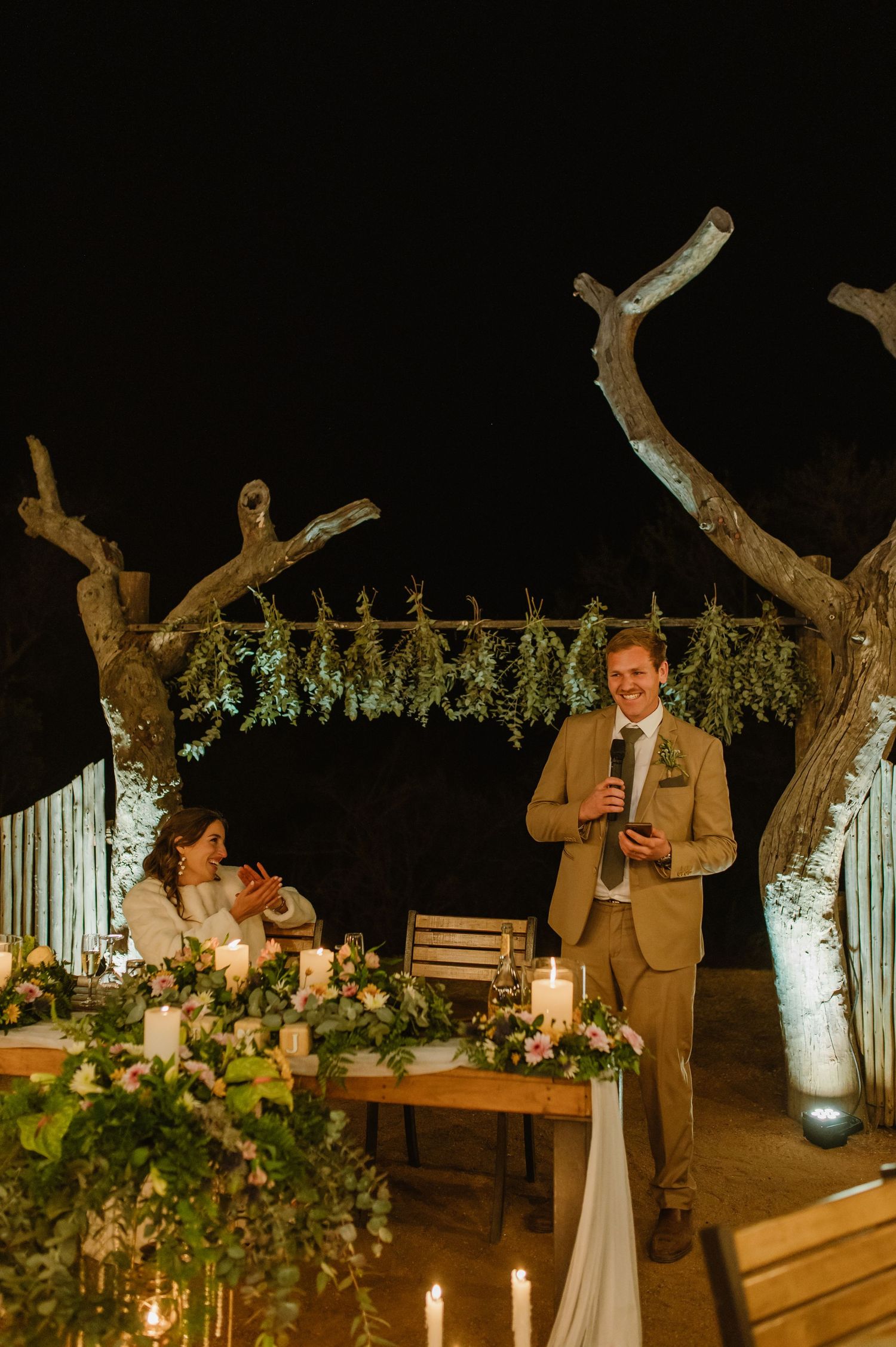 A wedding ceremony setup at night with string lights, white floral decorations, and rustic wooden elements.