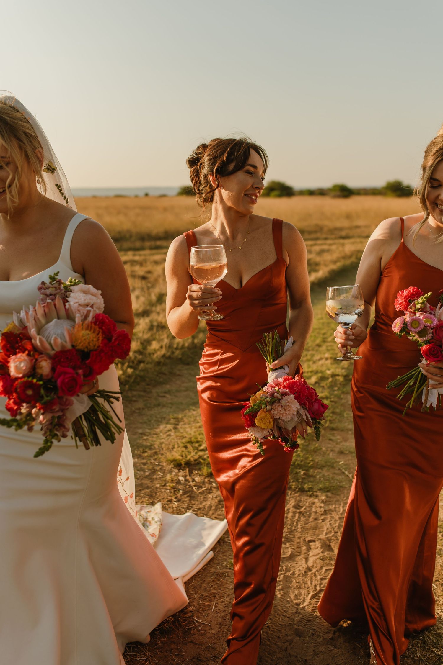 Bridesmaids in rust-colored dresses hold vibrant bouquets during golden hour photography.