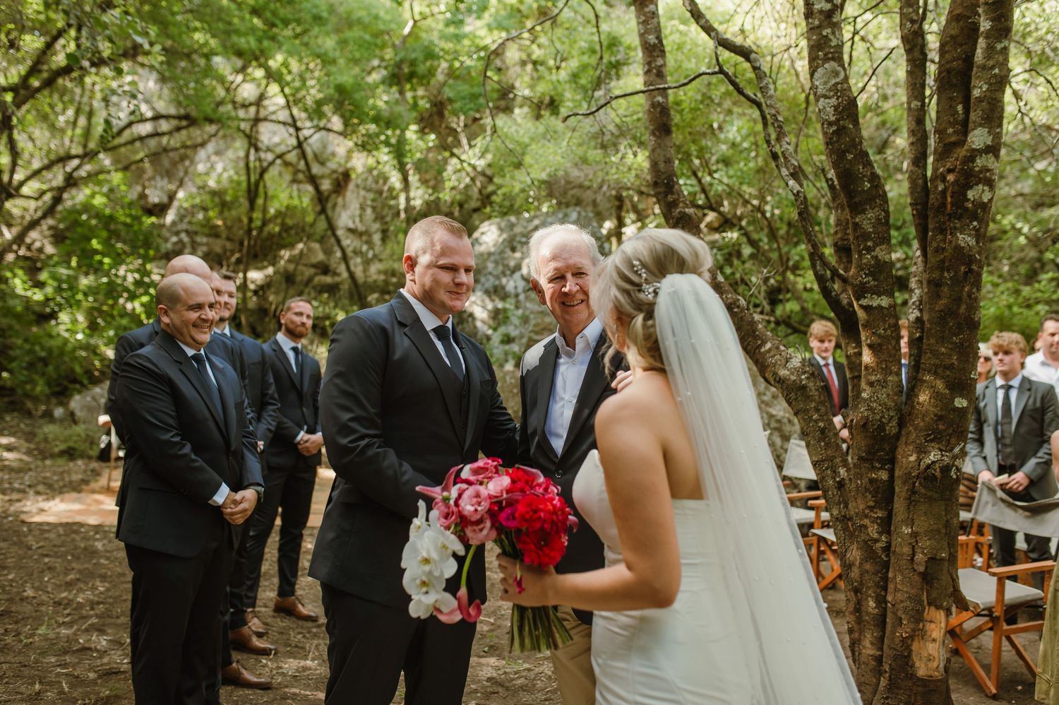 Wedding ceremony moment captured in a natural forest setting with red floral accents.