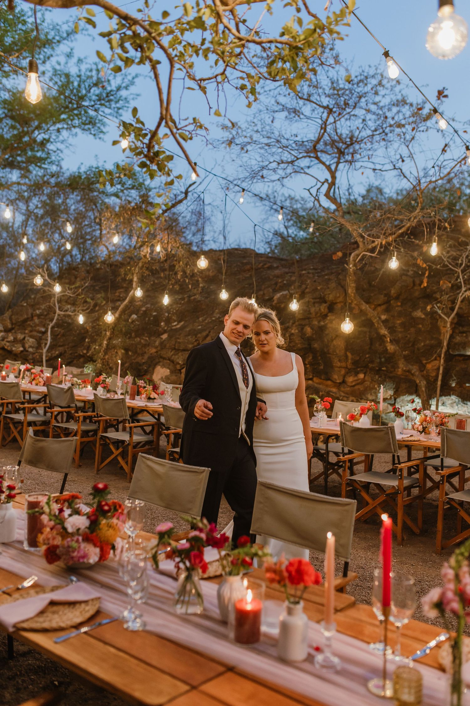An intimate outdoor wedding reception with hanging lights and red table decorations.