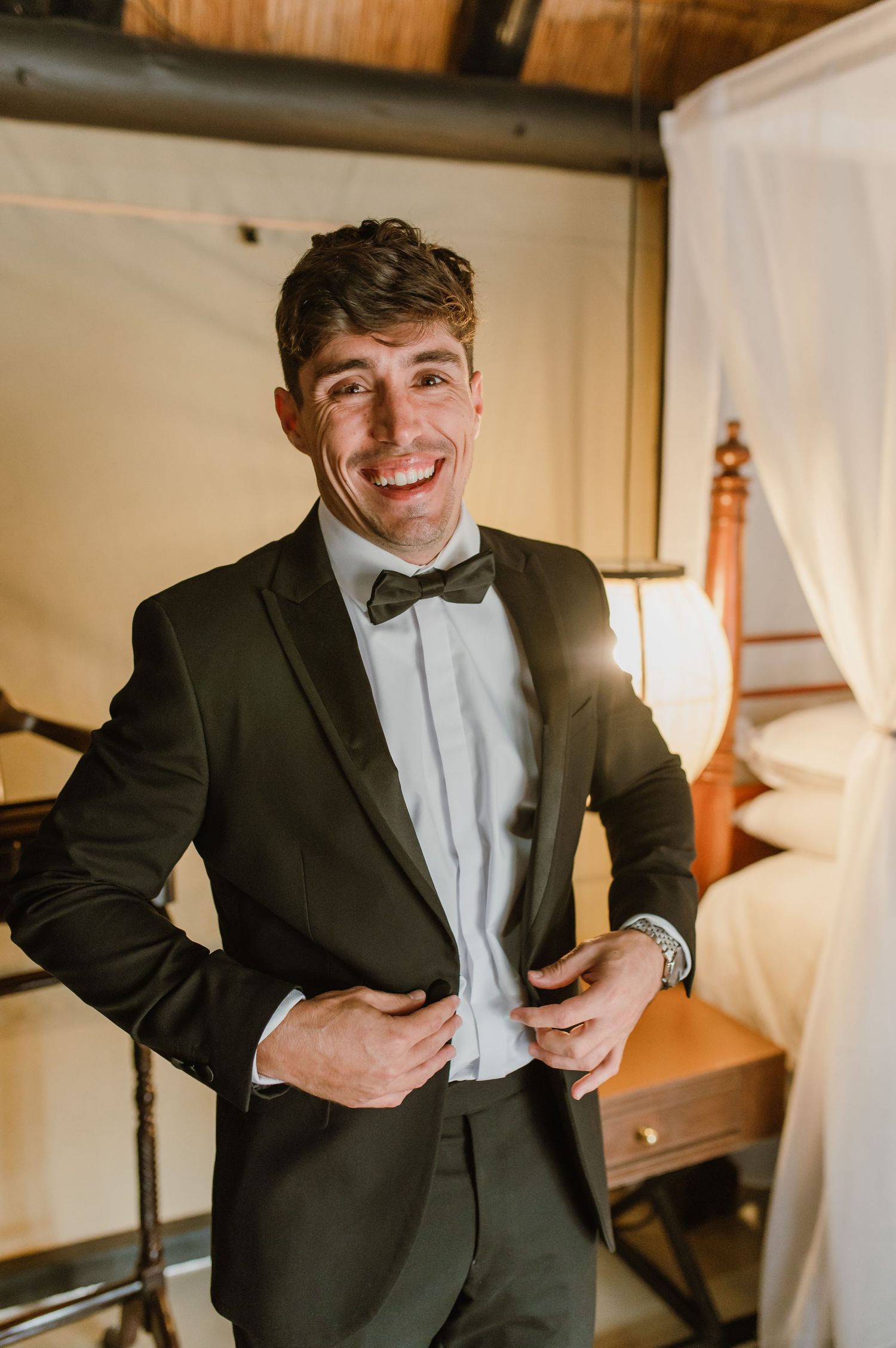Joyful portrait showing someone in a fitted black tuxedo adjusting their jacket indoors.