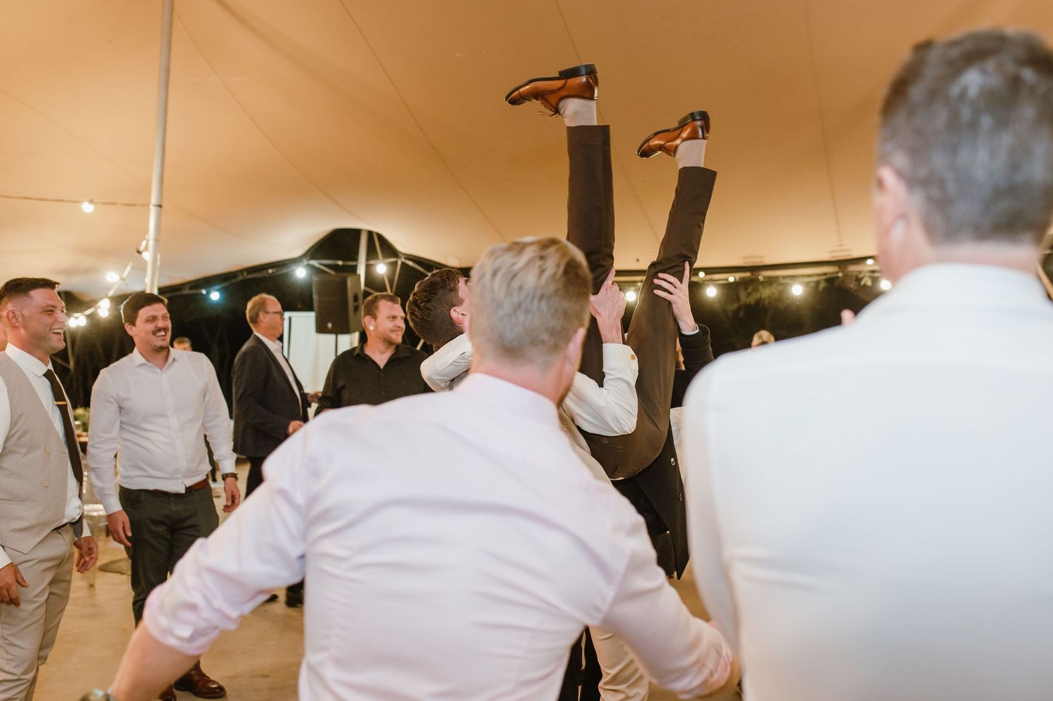 Guests perform acrobatic dance moves at a wedding reception as others watch and cheer.