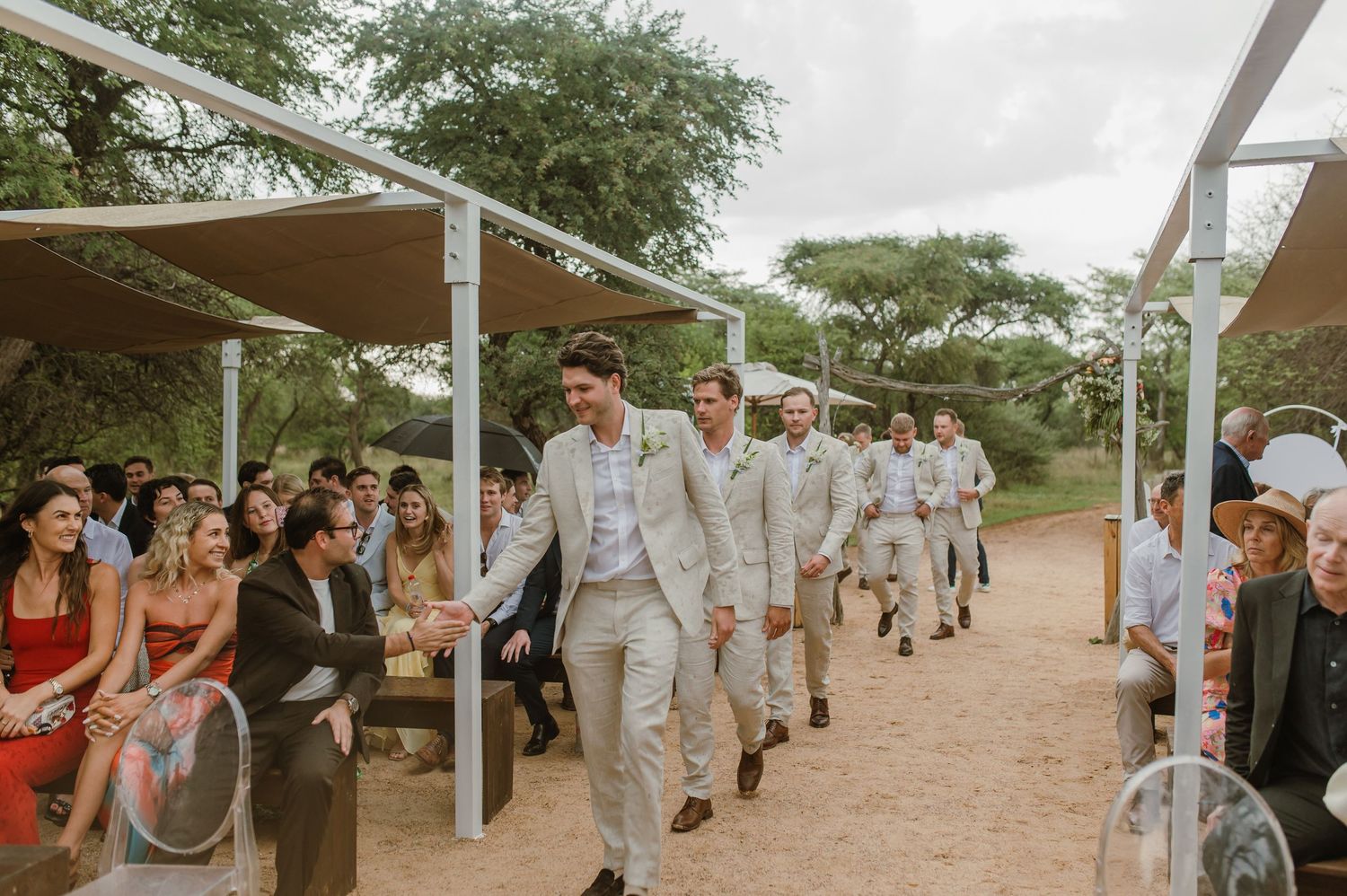 Wedding party members walk in procession on a dirt path under shade sails at an outdoor venue.