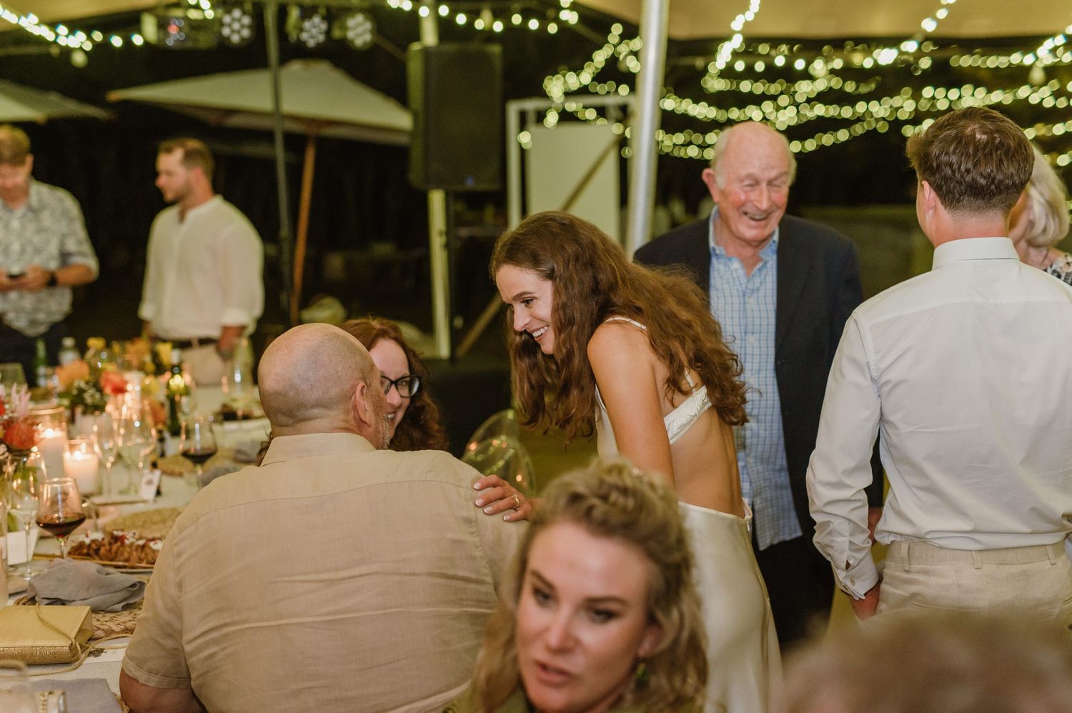 Wedding reception guests seated at long tables under string lights in an outdoor tent venue.