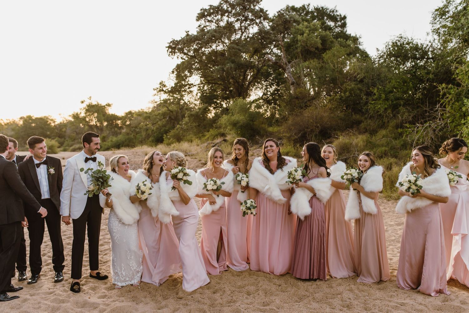 Bridal party in pink dresses and dark suits poses together on beach at sunset.