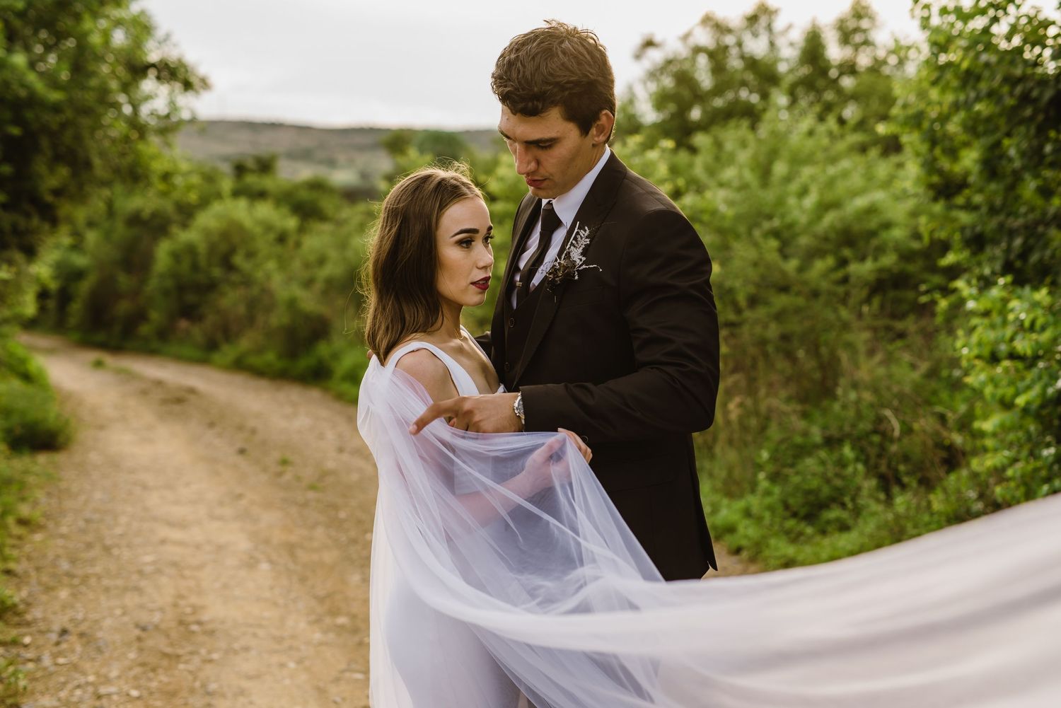 Wedding couple shares intimate moment with billowing white veil in natural outdoor setting.