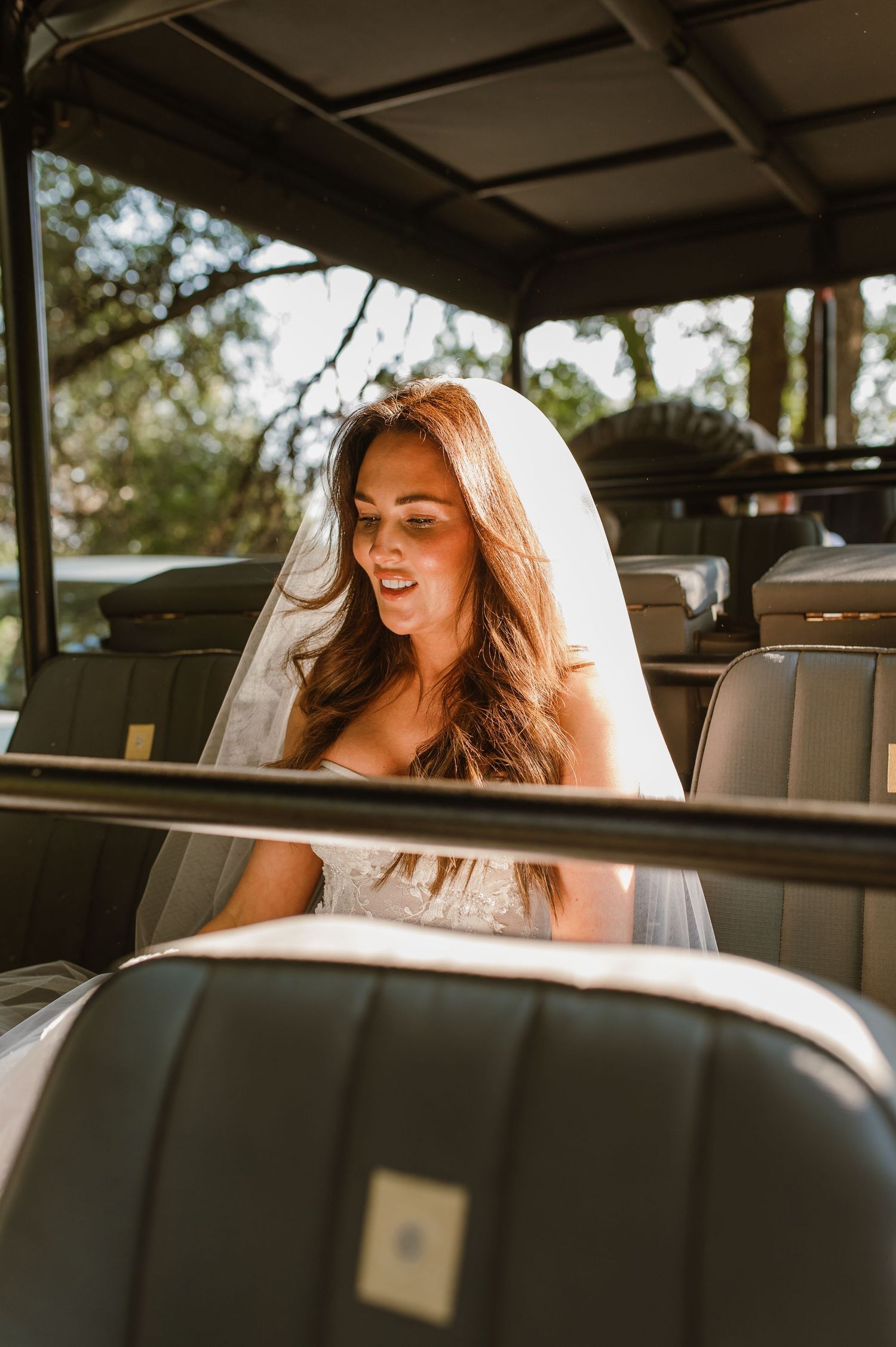 A radiant bride in a lace gown and veil sits inside a vintage vehicle bathed in golden light.
